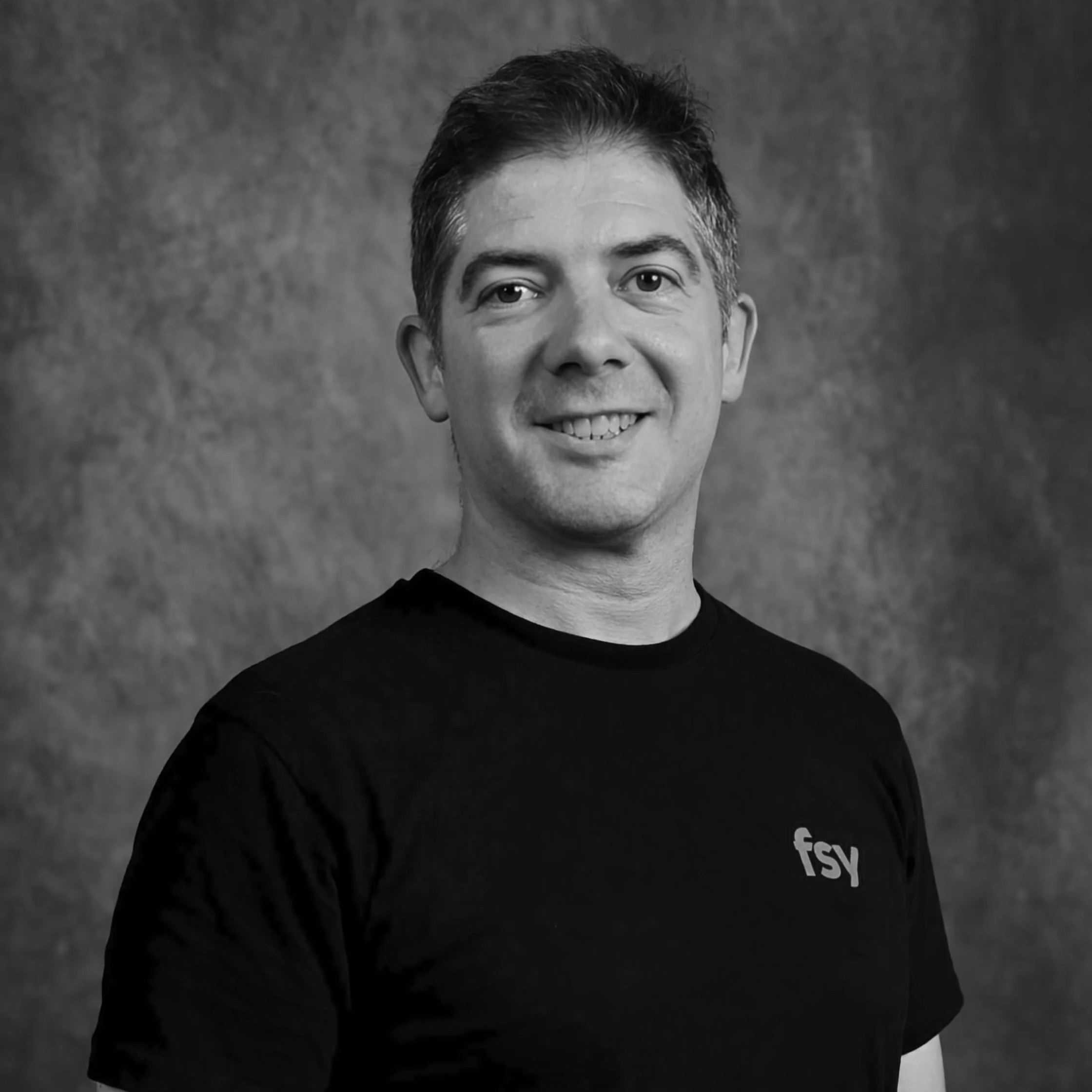 Portrait en noir et blanc d'un homme souriant, portant un t-shirt noir avec le logo "fsy".