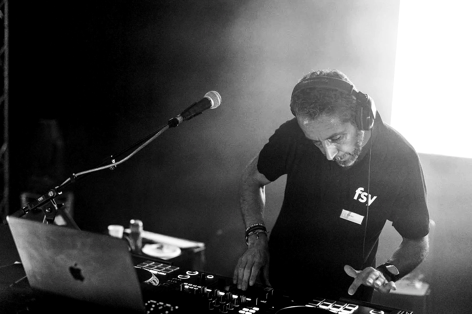 Un DJ en train de mixer avec des écouteurs, dans une ambiance sombre avec lumière forte derrière lui.