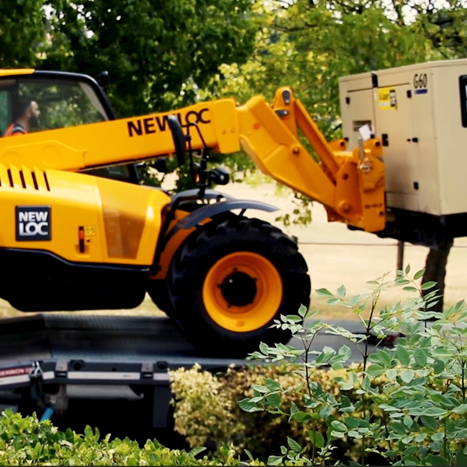 Une machine de construction jaune empilée sur une remorque, avec des arbres en arrière-plan et des buissons en premier plan.