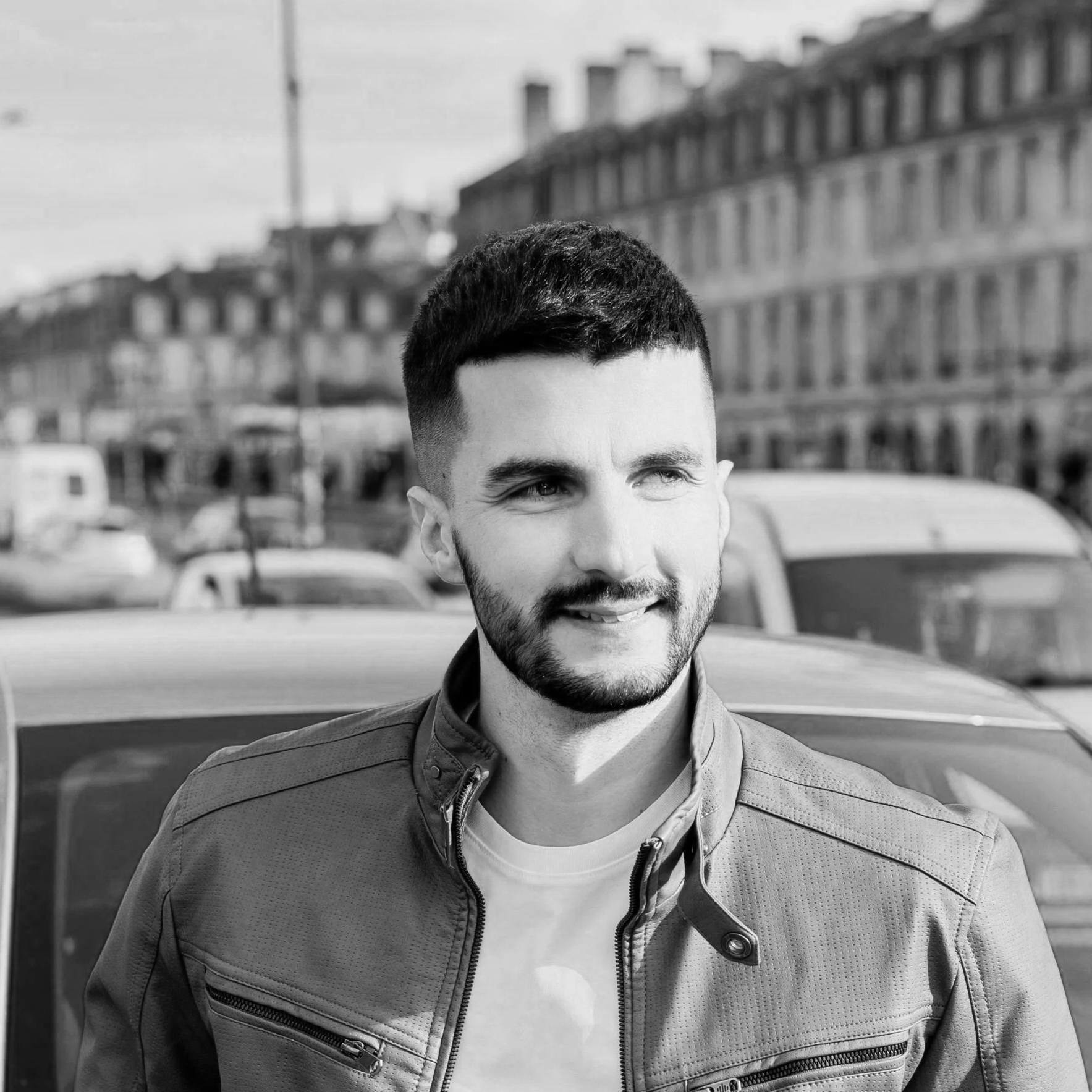 Un homme avec une barbe et des cheveux courts, portant une veste en jean, face vers la droite, en milieu urbain rencontré avec des voitures en arrière-plan.