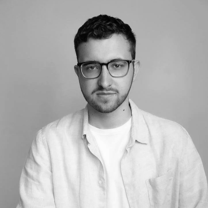 Un portrait en noir et blanc d'un jeune homme portant des lunettes, avec une barbe légère, portant une chemise légère sur un t-shirt, regardant vers l'objectif, sur un fond neutre.