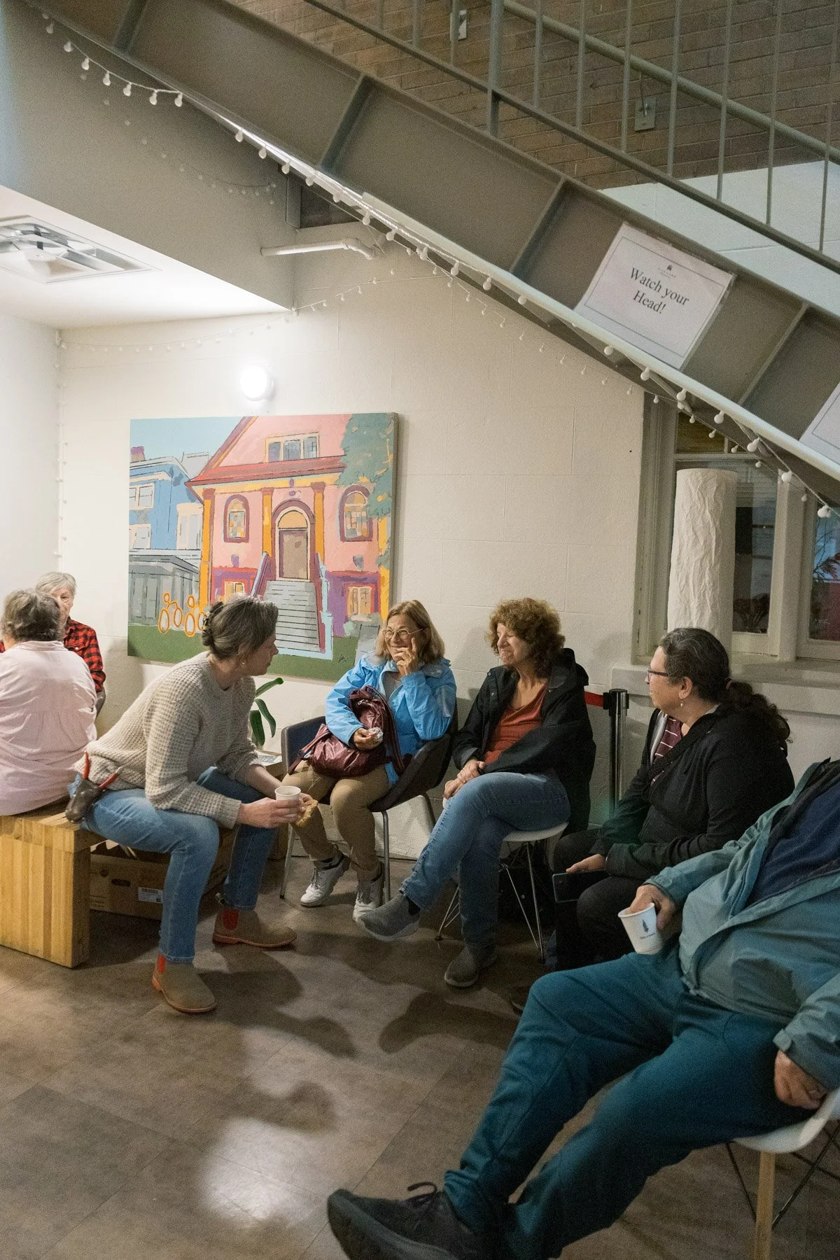 A group of people sitting and talking in a room with a painting of a building on the wall. Some are holding coffee cups.