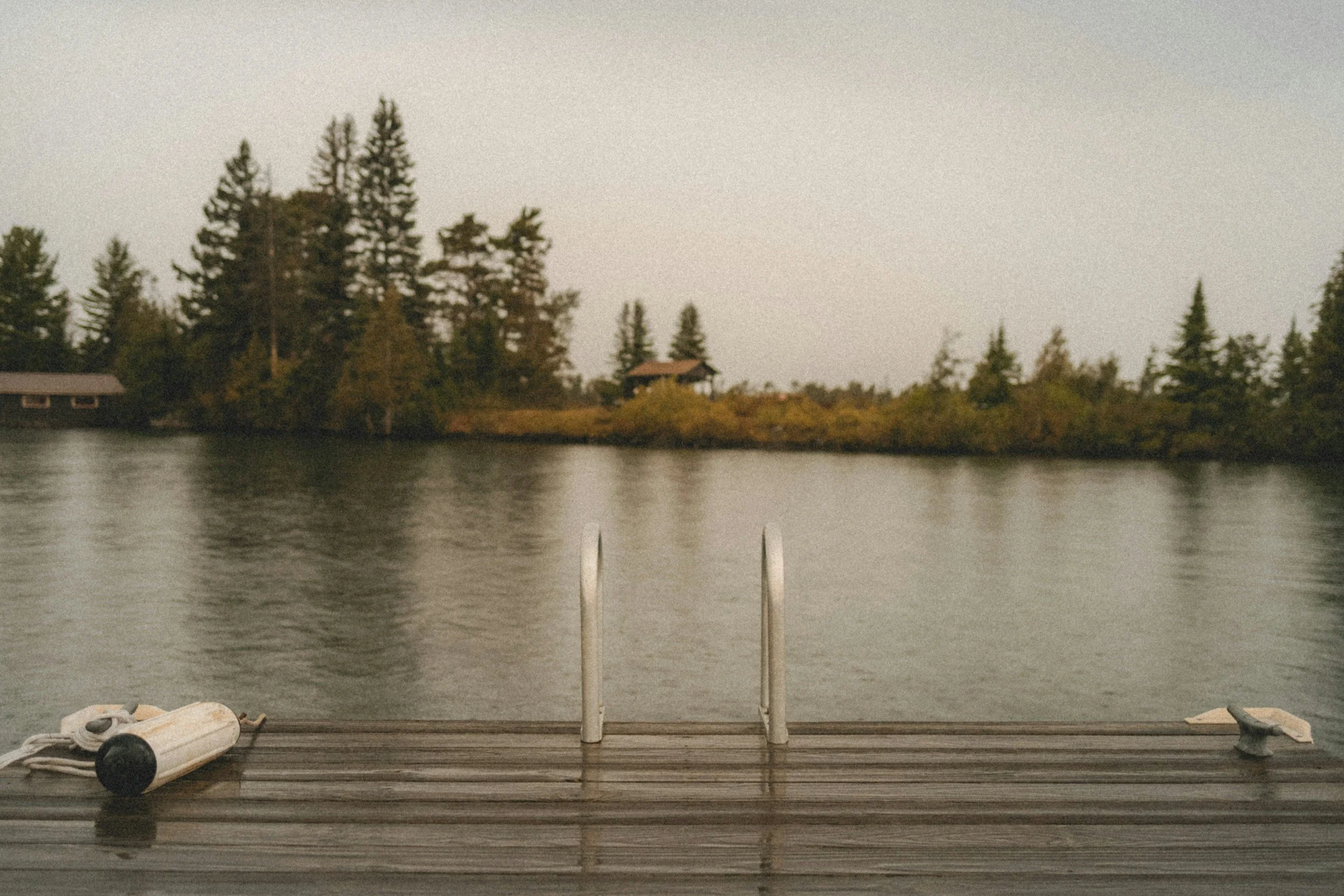 A view from a dock with a boat ladder leading into calm water, overlooking a wooded shoreline with trees and a house under a cloudy sky.