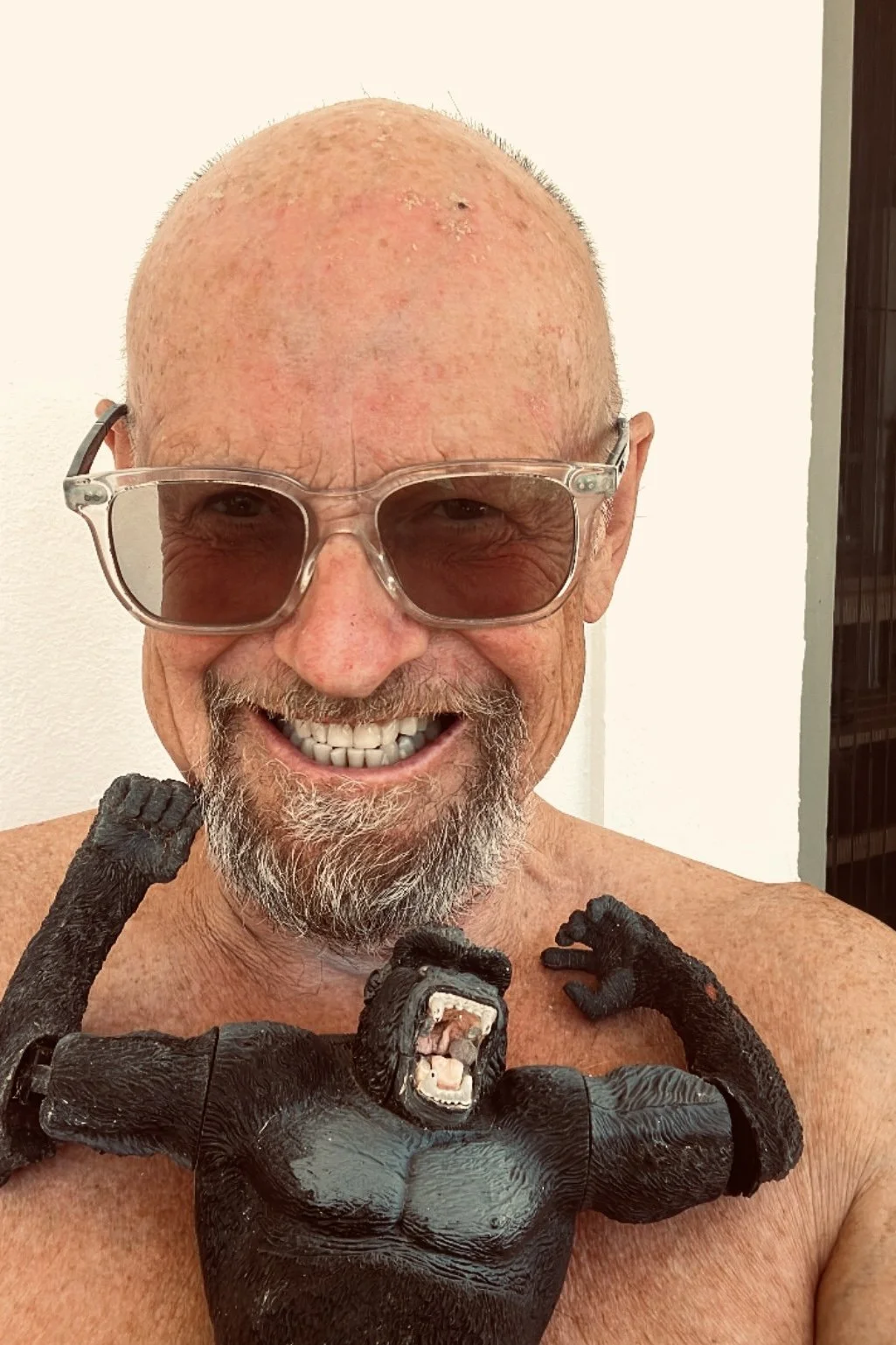 An older man with a bald head, glasses, and a goatee, smiling broadly. He is holding a large, black gorilla-shaped sculpture around his neck, which has its mouth open and arms raised.