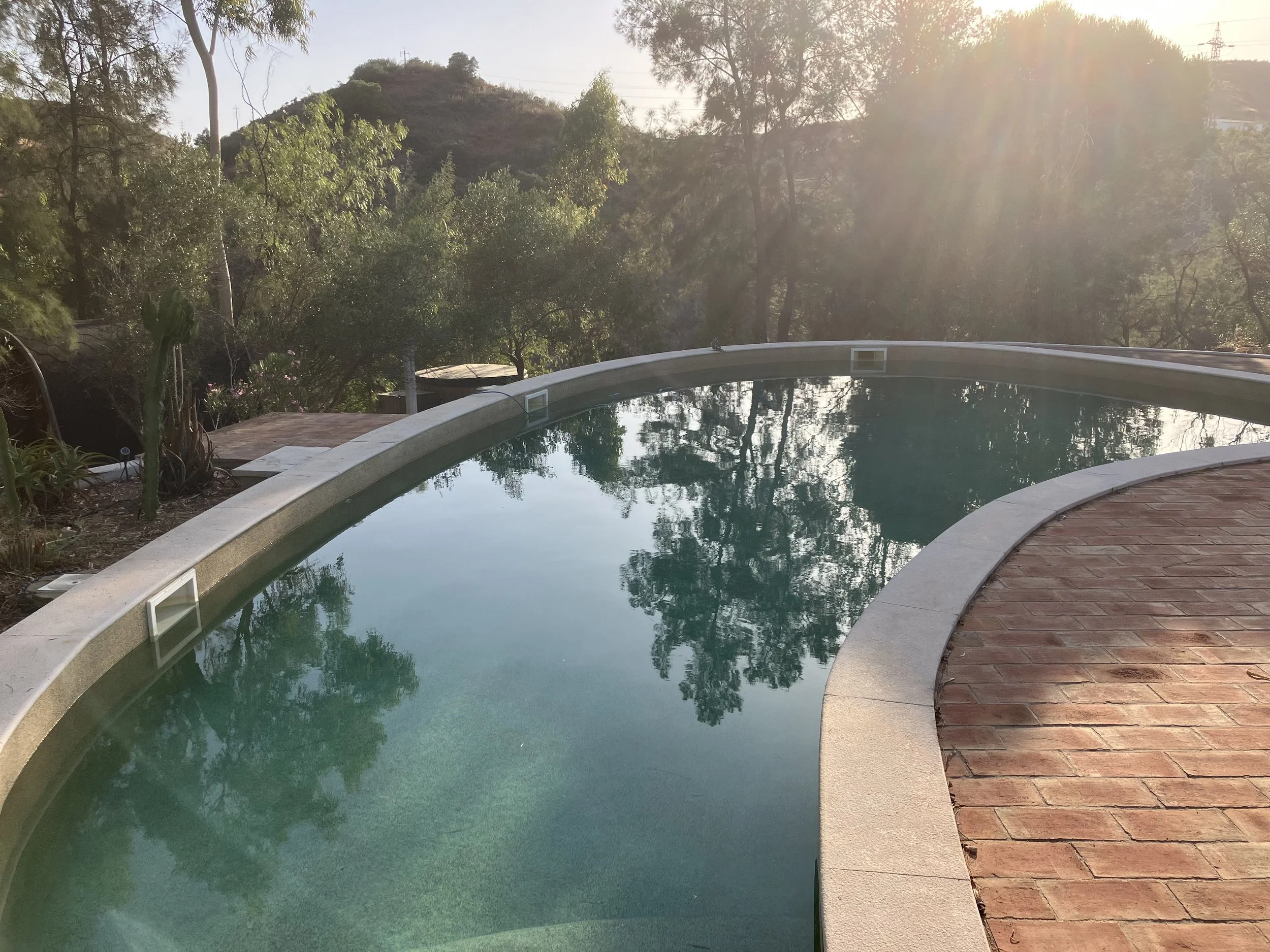 A swimming pool with a brick pathway on one side and trees and hills in the background, sunlight shining through the trees, creating a warm glow.