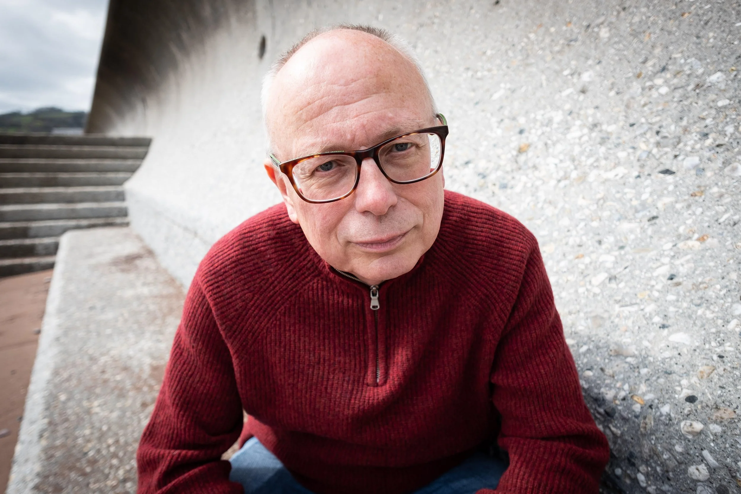 A man wearing glasses and a red zip-up sweater sitting on gravel outside, with concrete steps and a curved wall in the background.