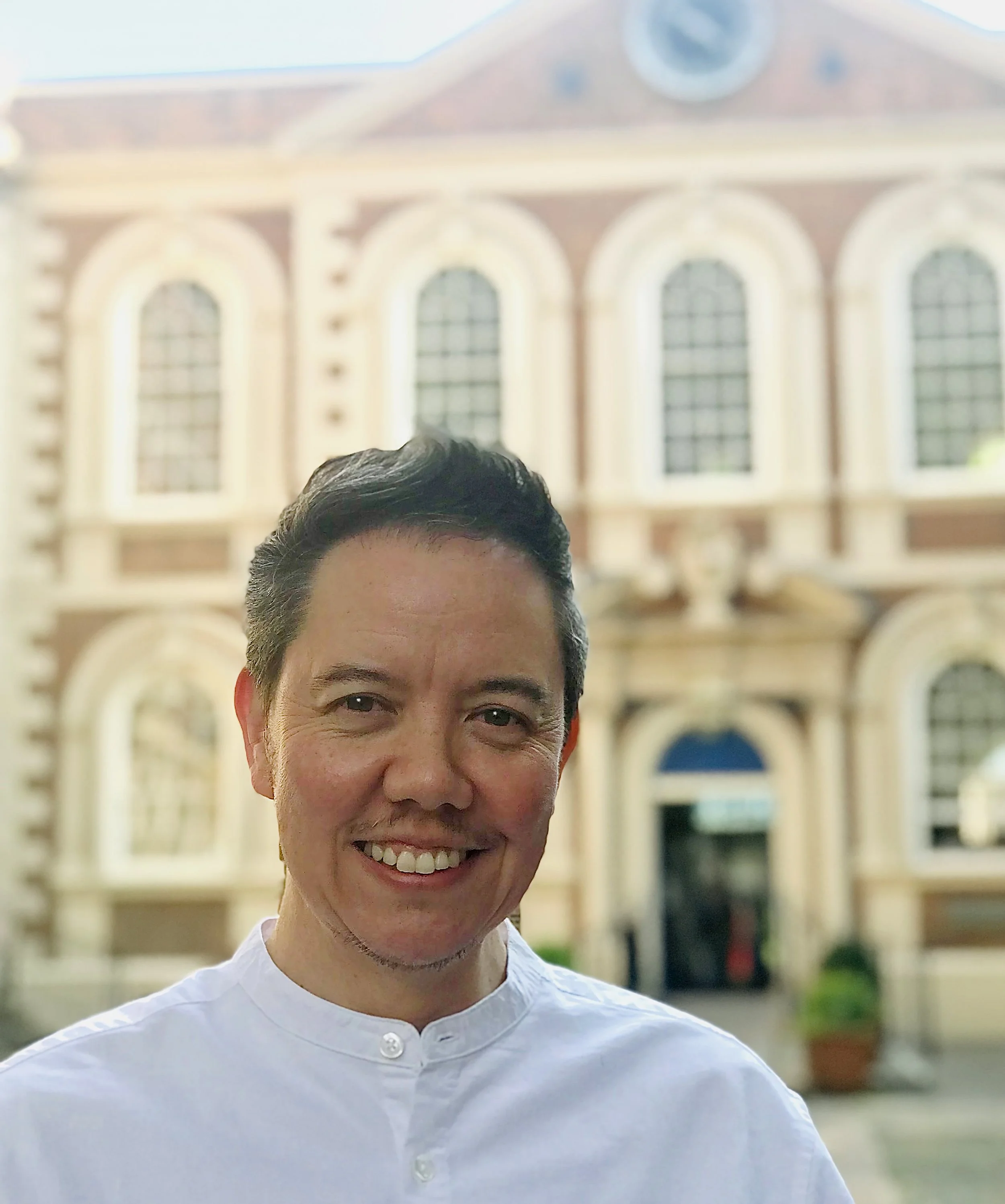 Smiling man with short black hair standing in front of a historic building with large arched windows and ornate architecture.