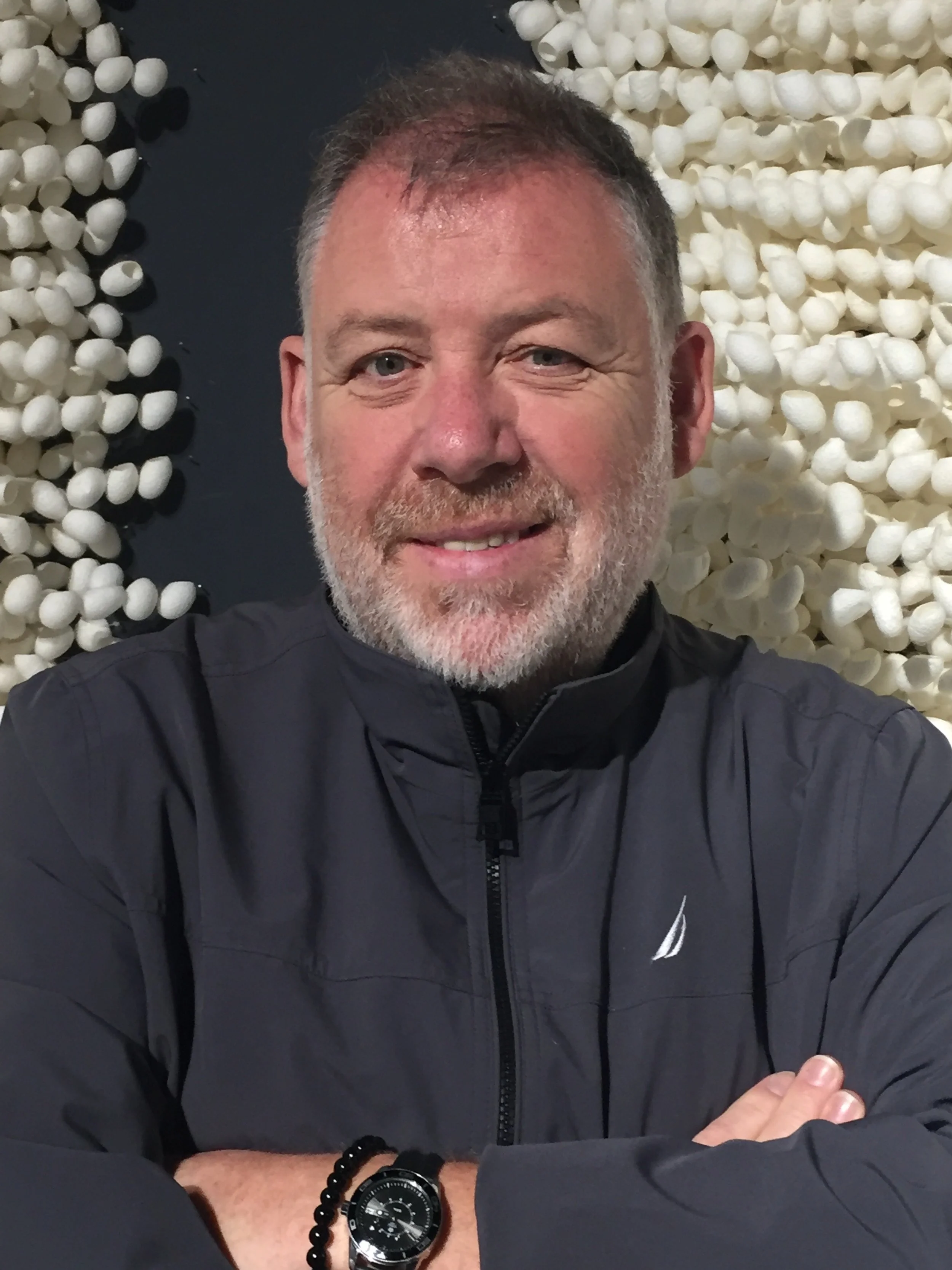 A middle-aged man with gray hair and beard smiling with arms crossed, wearing a black jacket and a watch, standing in front of a wall decorated with white rounded objects.