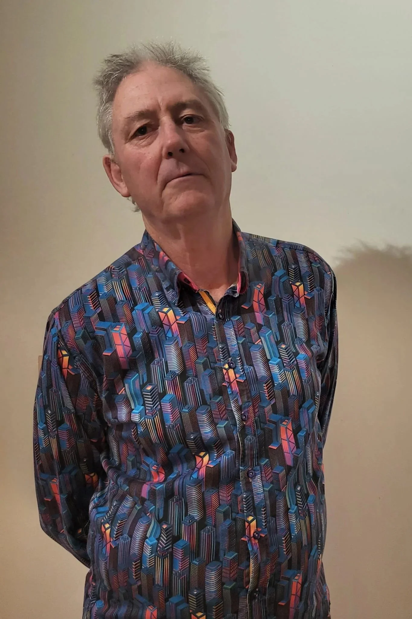 A man with gray, messy hair and a serious expression, wearing a colorful, patterned long-sleeve shirt, standing against a plain white wall.