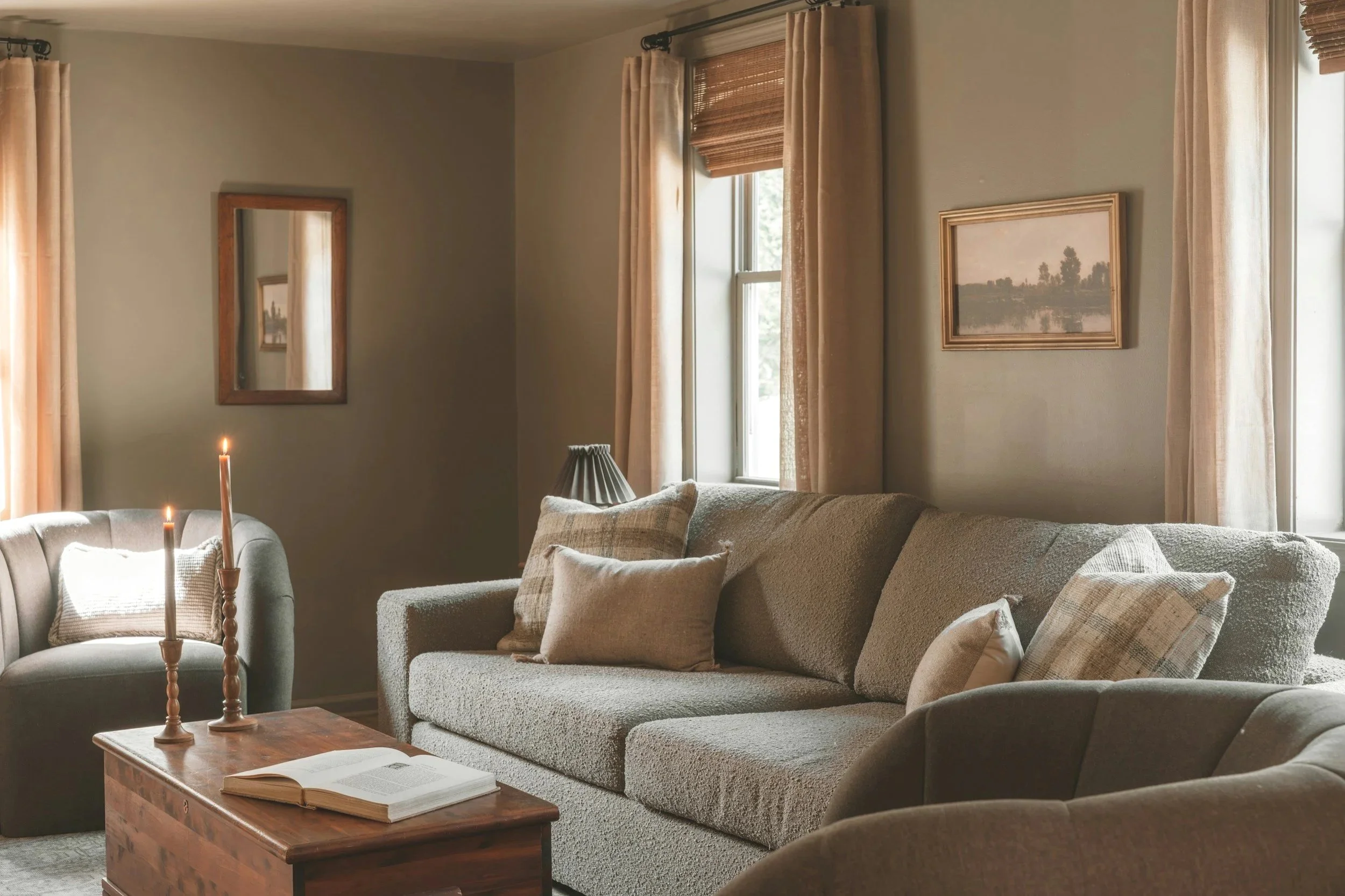 Cozy living room with beige sofas, candles, open book on wooden coffee table, framed landscape painting, and windows with curtains and bamboo blinds.