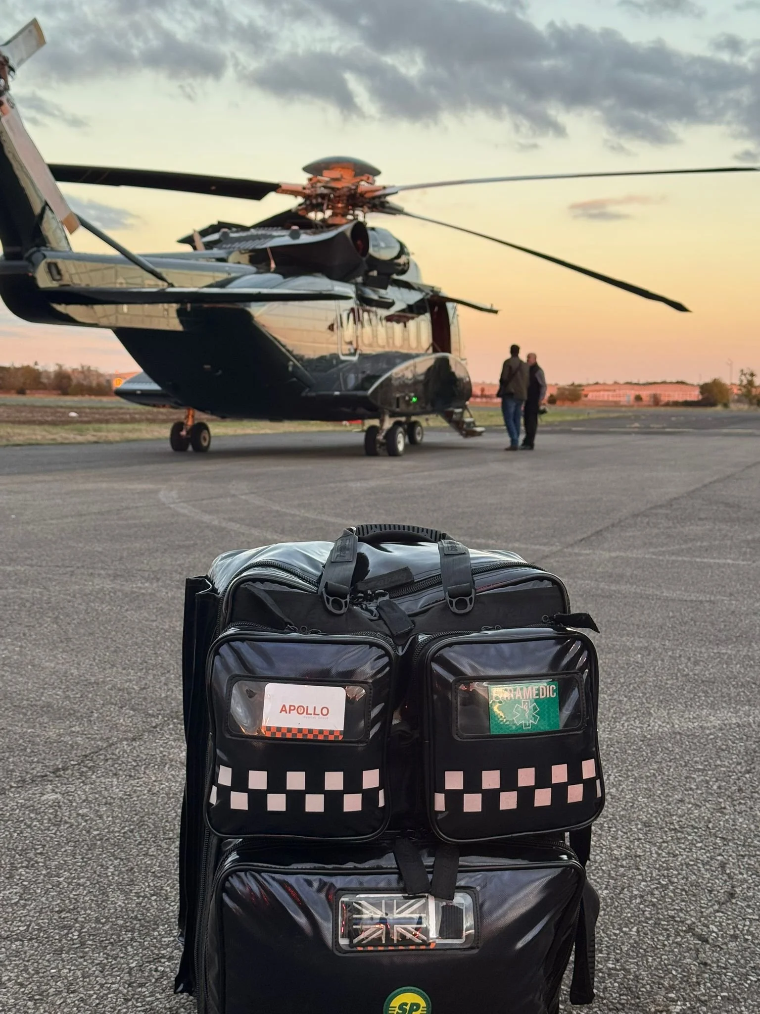 A black emergency medical bag with patches labelled 'Apollo' and 'Paramedic' on a tarmac, with a special escort helicopter and two people in the background at sunset.