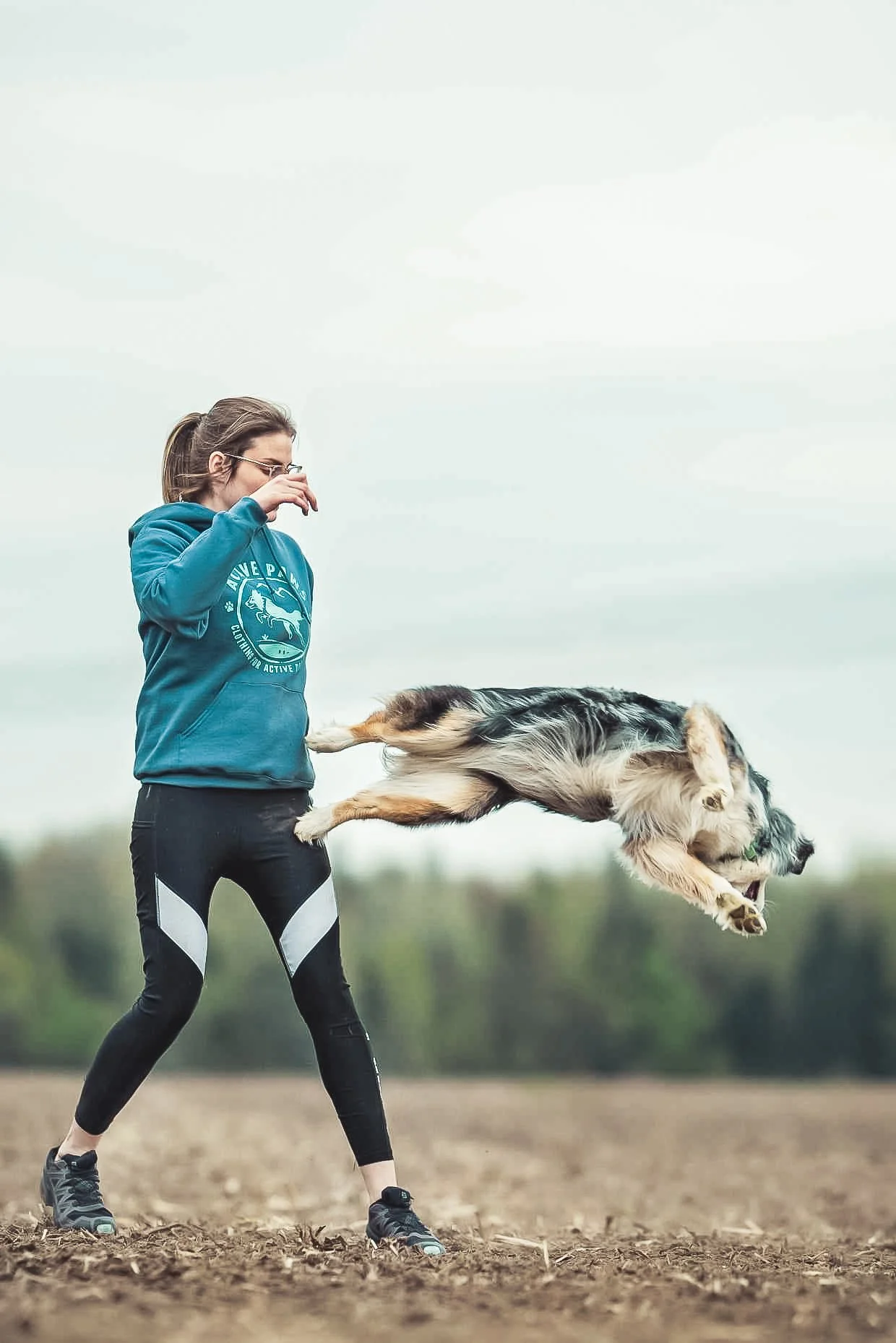 Une femme lance un chien en plein air, le chien est en mouvement dans un espace ouvert et herbeux, avec un ciel nuageux en arrière-plan.