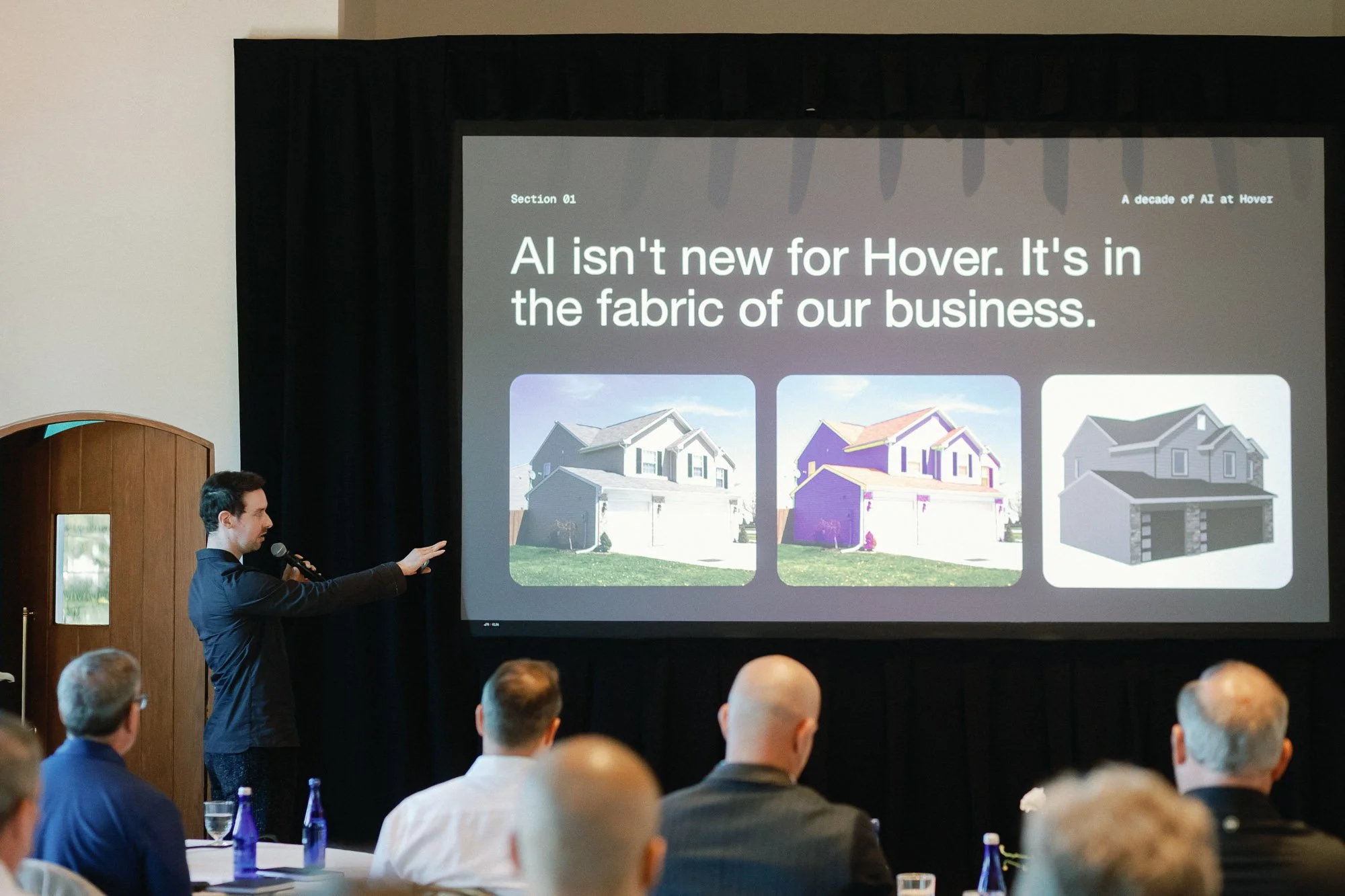 A man in a dark suit giving a presentation with a microphone in front of a projection screen showing images of houses and text about AI in the housing industry, in front of an audience of seated and standing people.