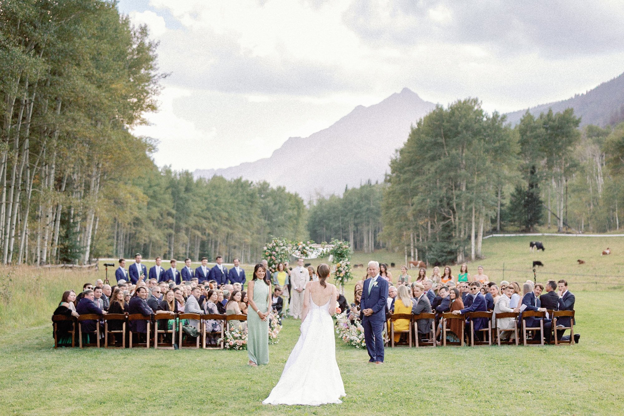 A wedding ceremony taking place outdoors on a green field surrounded by trees and mountains, with guests seated in chairs and a bride and groom standing at the altar with an officiant.