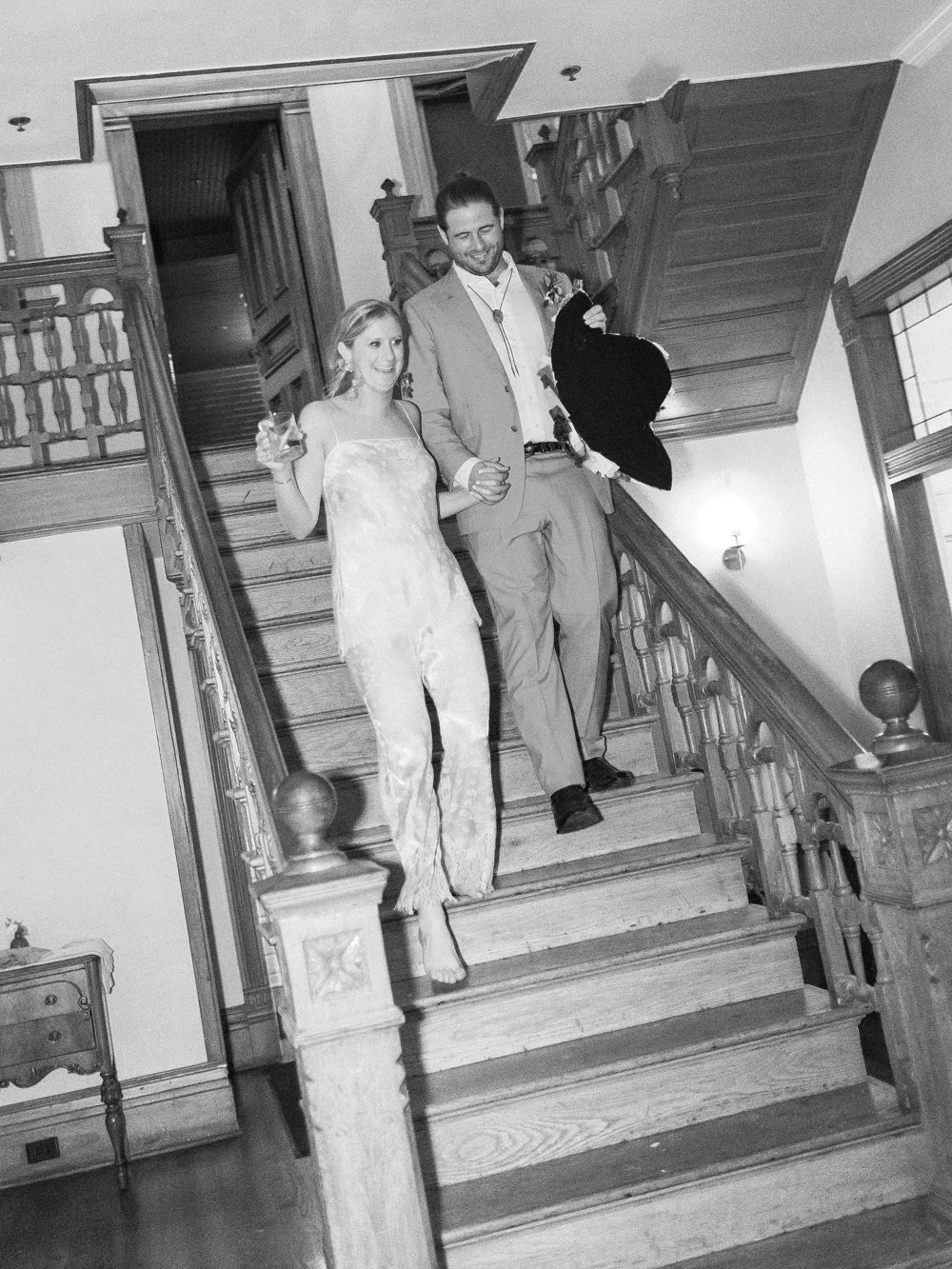 A couple walking down Mattie's wooden staircase at a wedding reception, holding drinks and smiling.