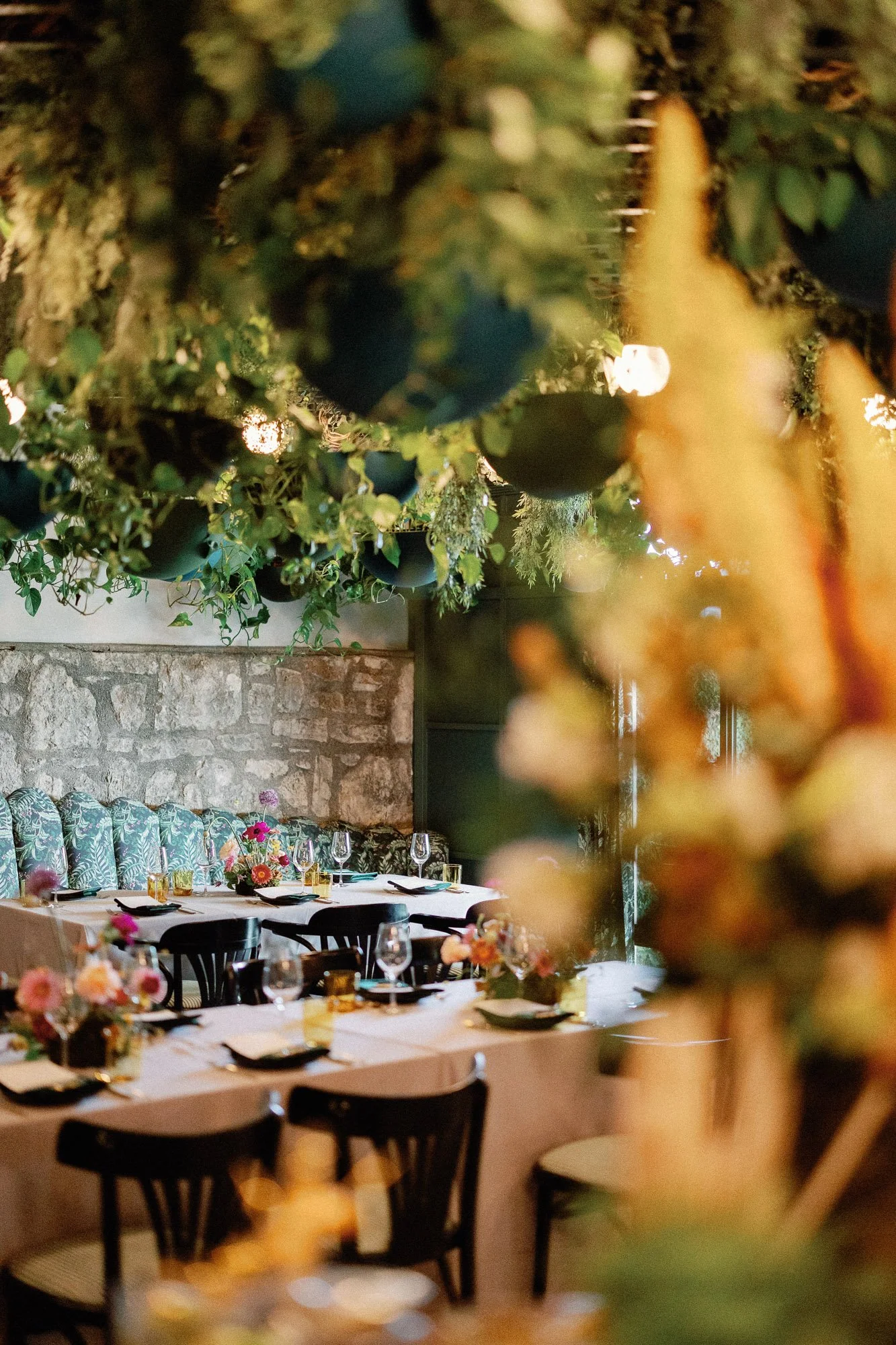 A dining area decorated for a celebration or event with a long table set with plates, glasses, and floral centerpieces. The room features a stone wall and lush greenery overhead, creating an elegant and inviting atmosphere.