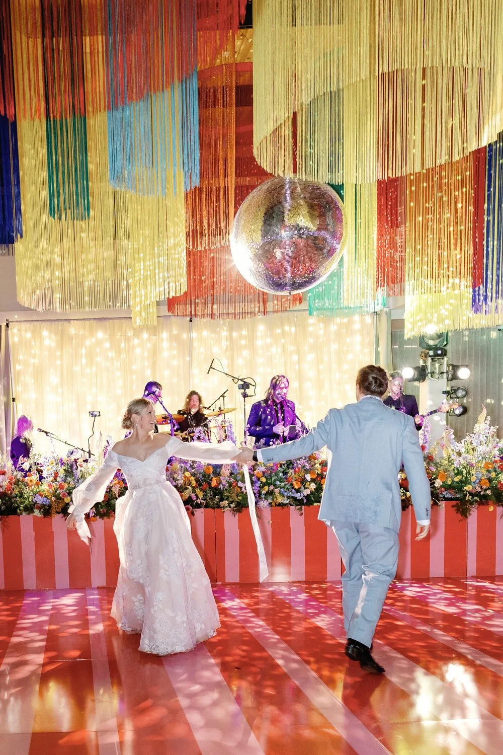 A bride and groom are dancing at their wedding reception in front of a live band, with colorful hanging decorations and a large disco ball overhead, and flower arrangements along the stage background.