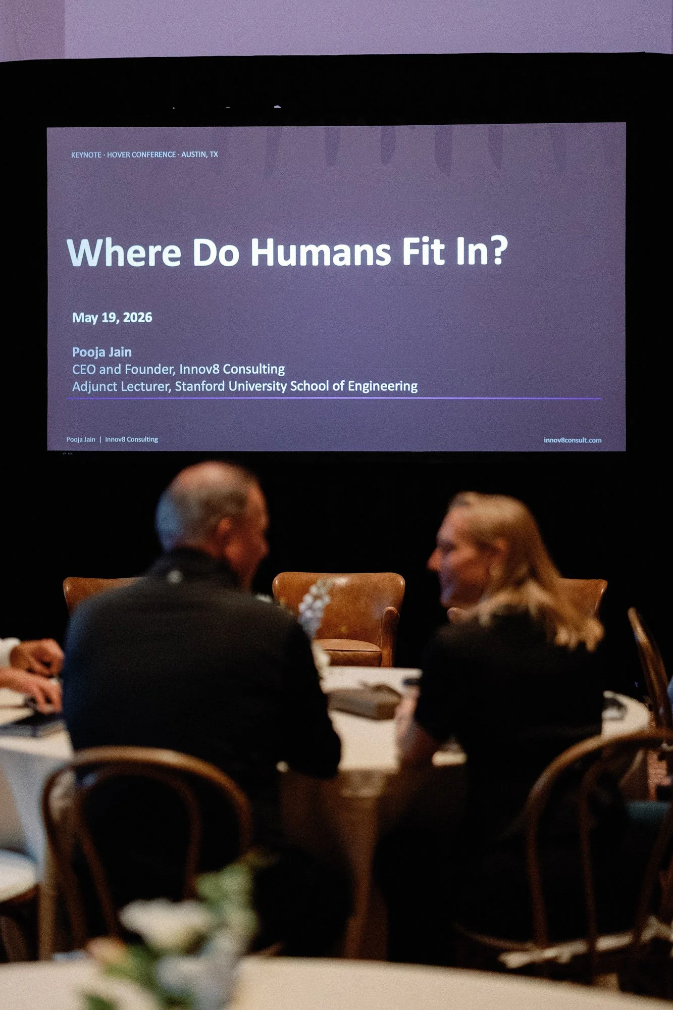 A presentation slide titled 'Where Do Humans Fit In?' displayed on a screen at a conference in Austin, Texas on May 19, 2026, with several people seated at a table in the foreground.