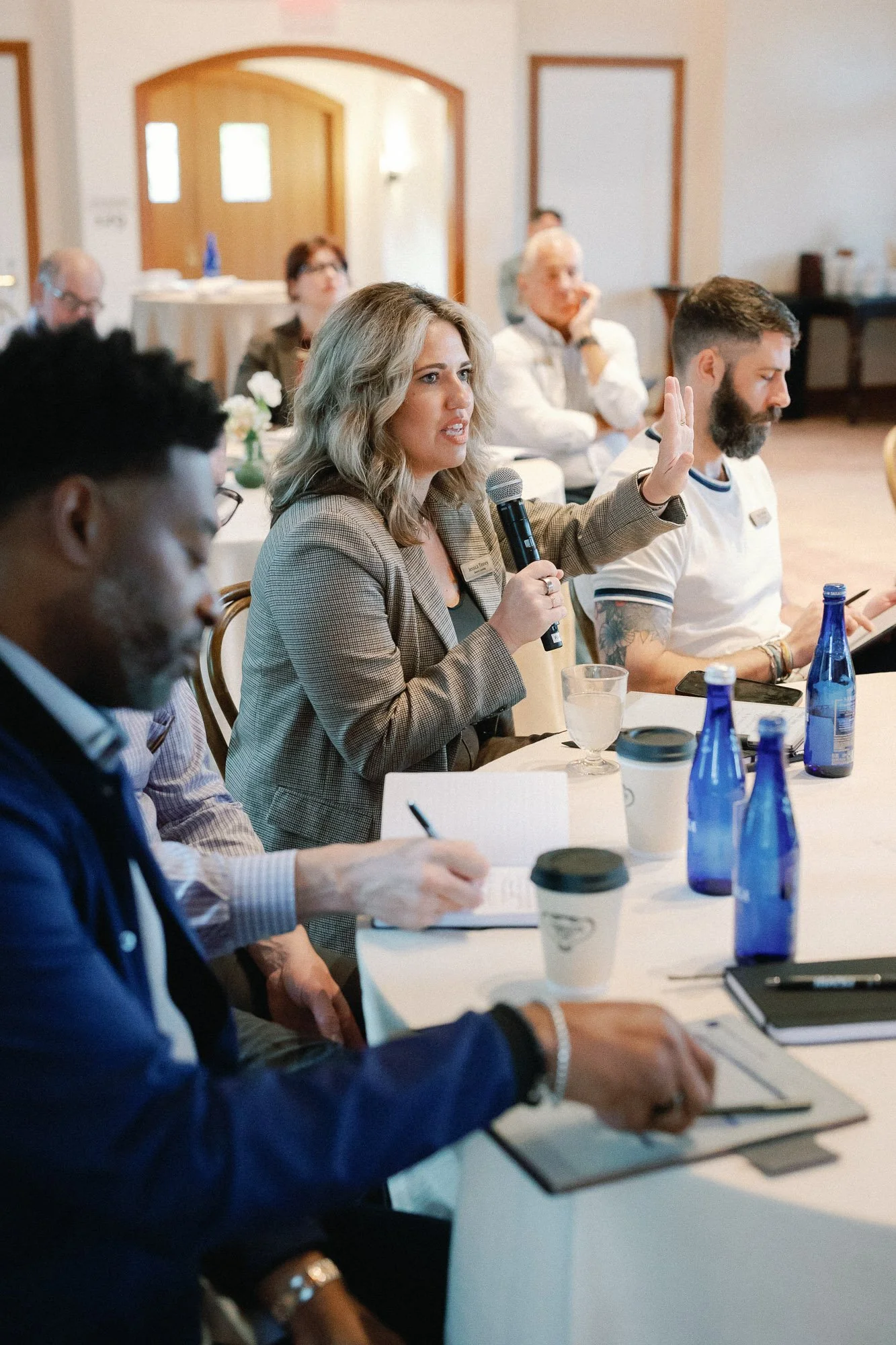 A woman is speaking into a microphone, gesturing with her hand during a meeting or conference. Several other people are seated around the table, listening attentively. There are water bottles and coffee cups on the table.