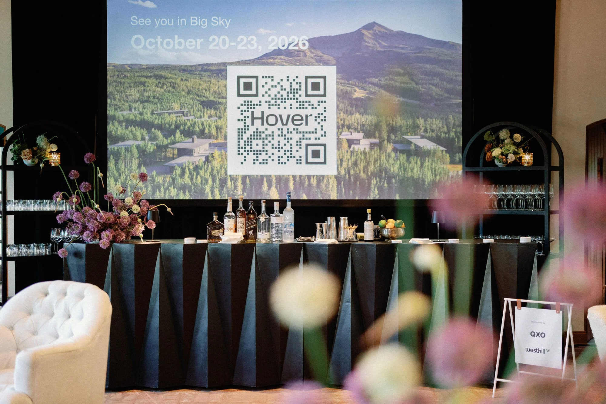 Large screen displaying an advertisement for an event with the phrase "See you in Big Sky," scheduled for October 20-23, 2026, featuring a QR code labeled "Hover." The room has a bar setup with liquor bottles, glassware, and floral arrangements, with