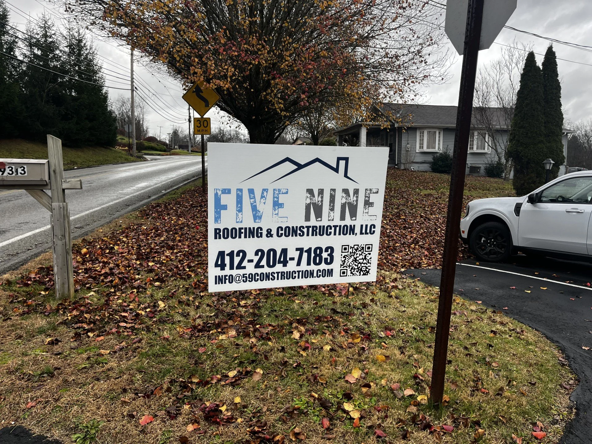 A sign on a grassy area with fallen leaves, advertising Five Nine Roofing & Construction, LLC, with a mountain outline and a house roof illustration. Contact number and email are displayed along with a QR code.