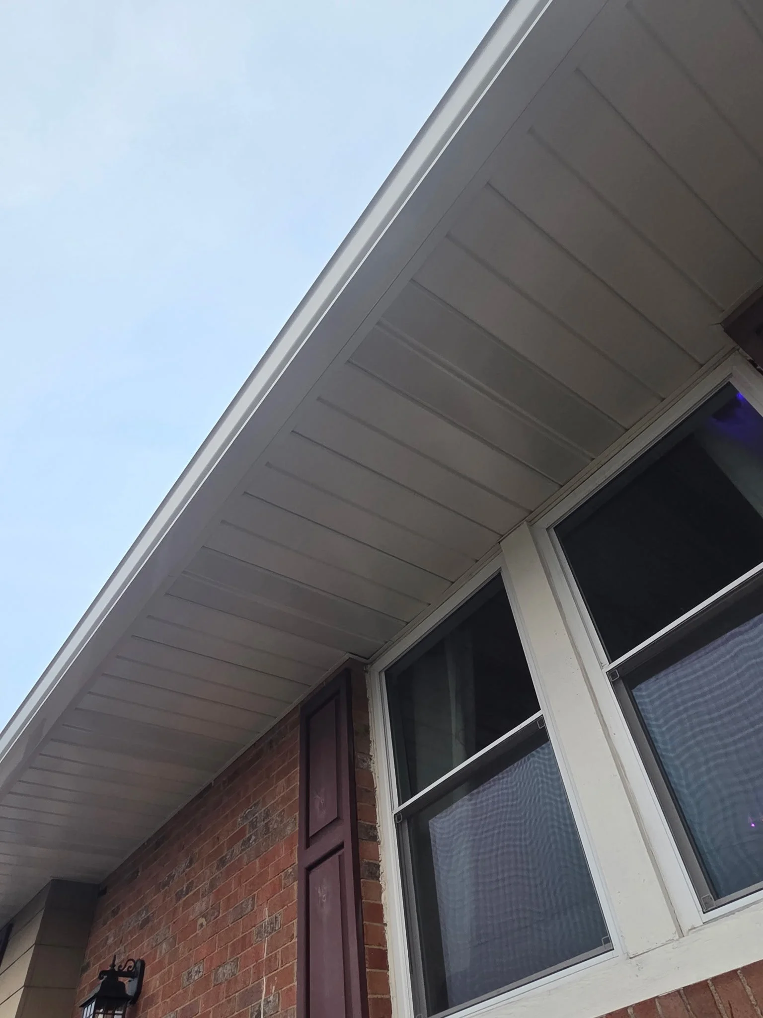 Close-up of a house exterior showing a brick wall, white window frames, and beige siding, with a clear sky in the background.