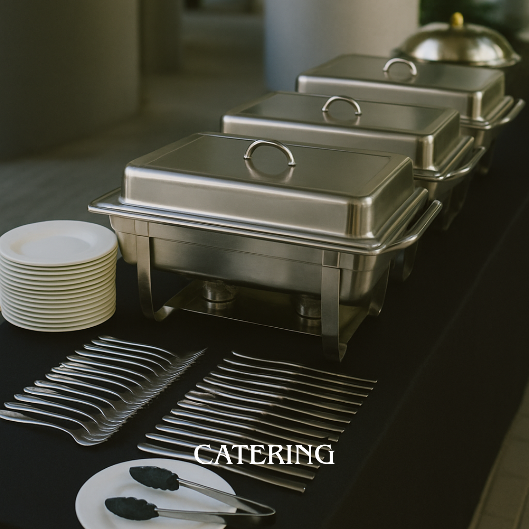 A buffet table with three silver chafing dishes covered with lids, a stack of small white plates, several pairs of silver tongs, and a pair of black tongs on a small white plate. The word 'CATERING' is overlaid on the image.