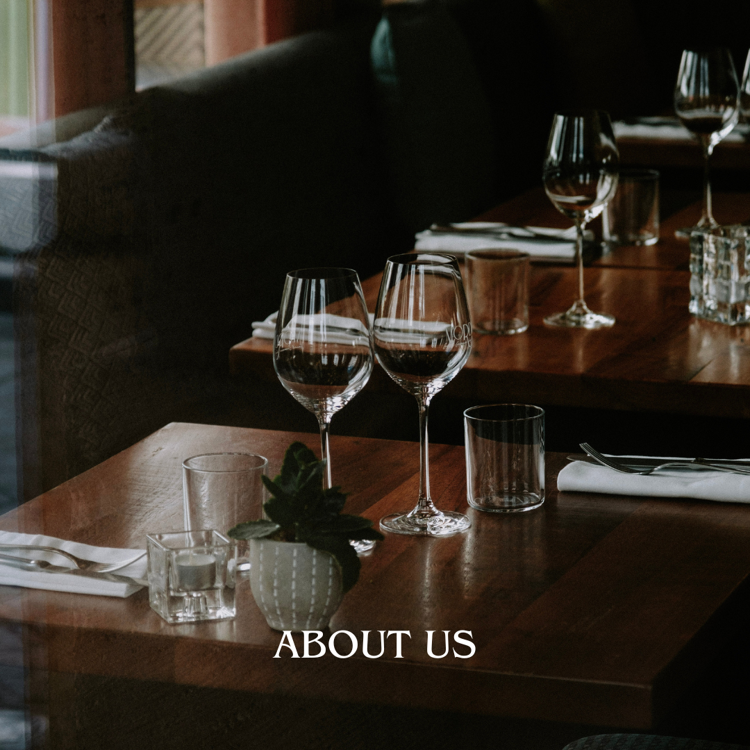 Empty restaurant table with wine glasses, water glasses, a small potted plant, and white napkins, with the text 'ABOUT US' at the bottom.