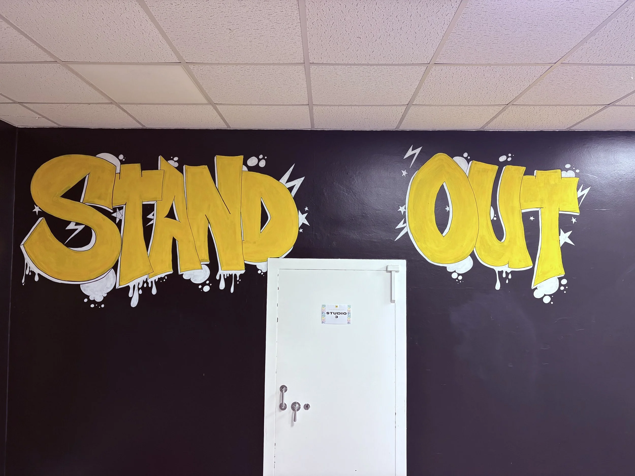 Graffiti-style wall art with the words 'STAND OUT' in bold yellow letters, decorated with white star and lightning bolt accents on a black wall above a white door.
