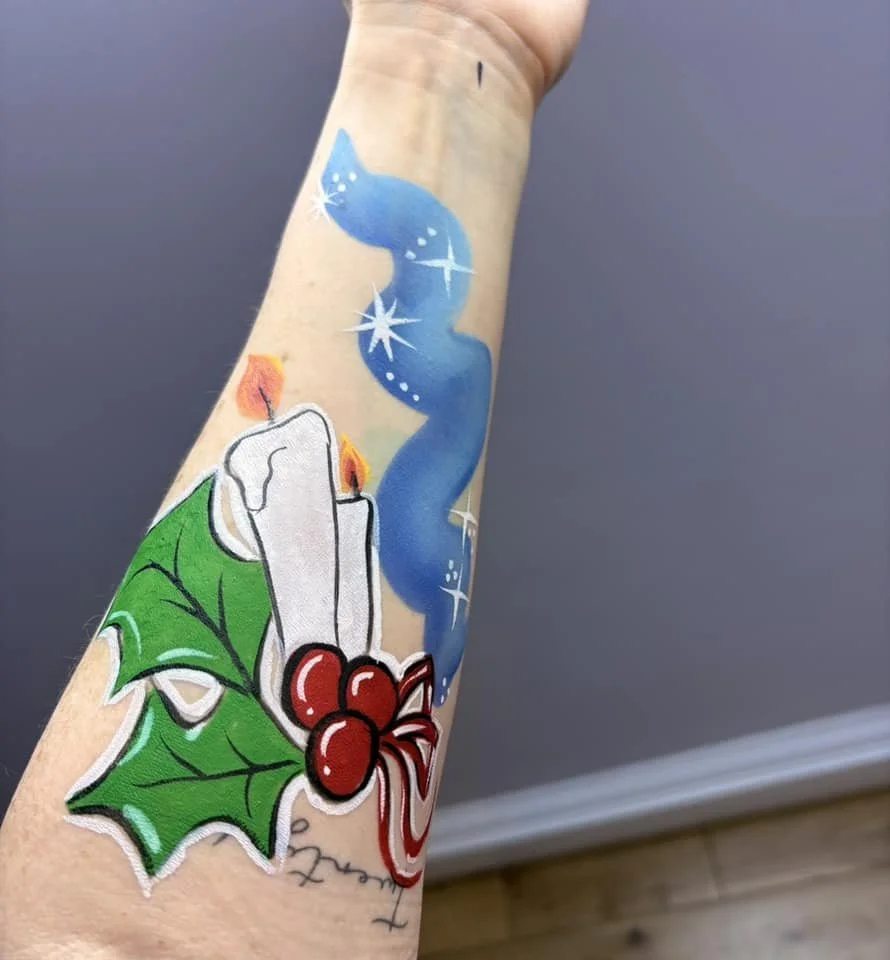 The image shows holiday-themed face paint on a forearm. There is a white candle with holly leaves and berries. There is also a blue, swirling, starry design extending above the candle.
