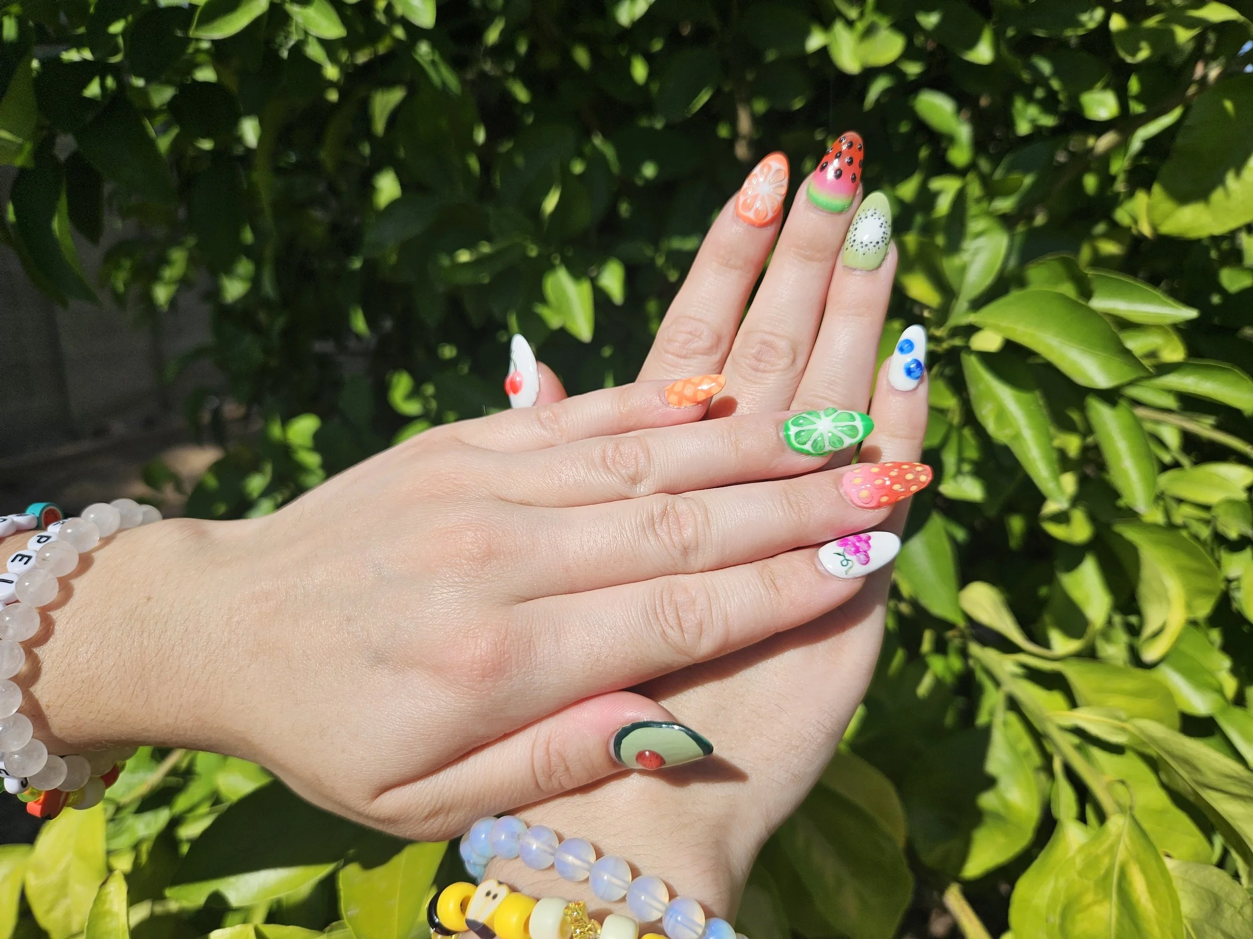 Close-up of two hands with colorful, fruit-themed nail art, against a green leafy background. The hands are adorned with beaded bracelets.