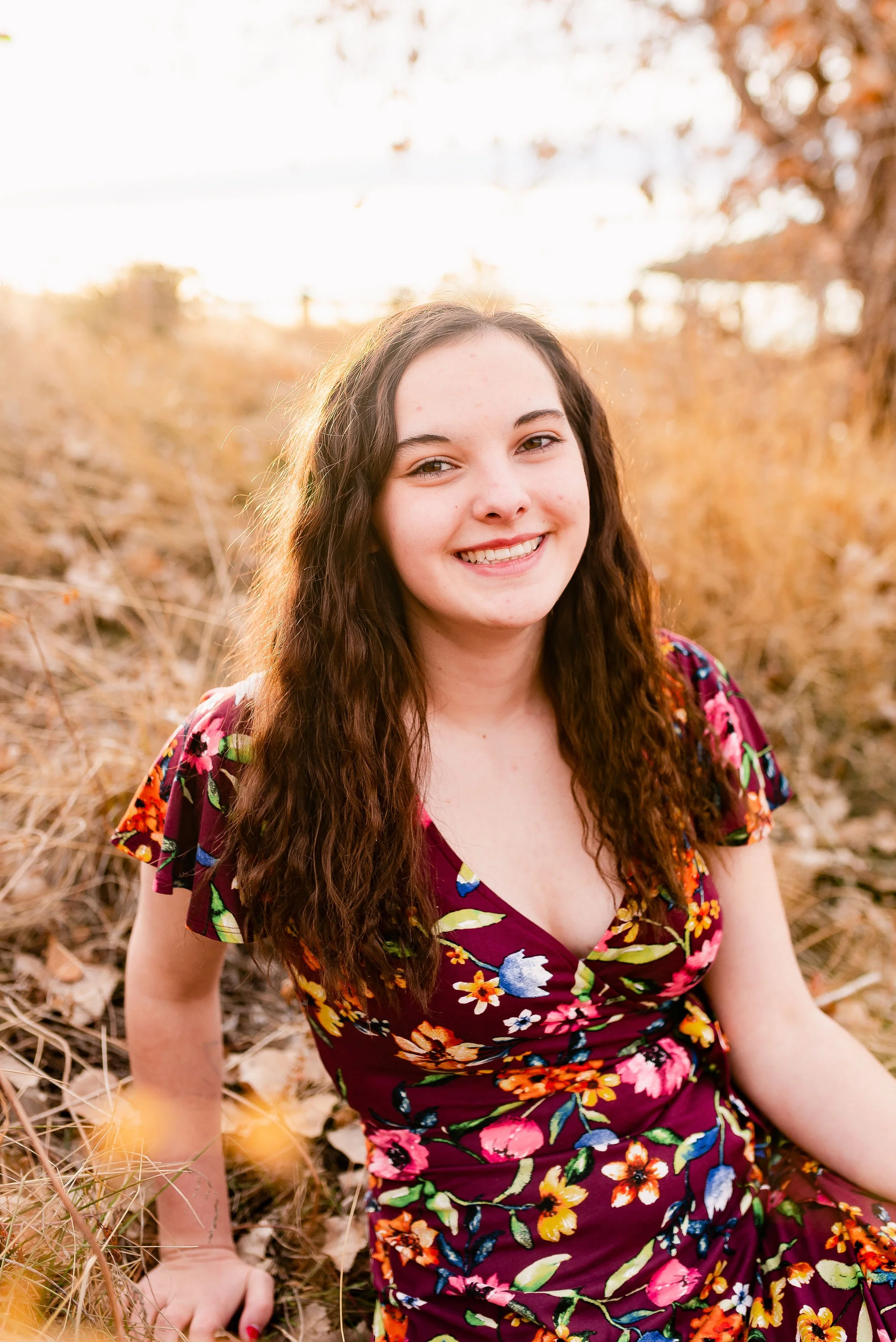 A young woman with long wavy brown hair smiling outdoors in a field of dried grass and leaves during sunset, wearing a colorful floral dress.