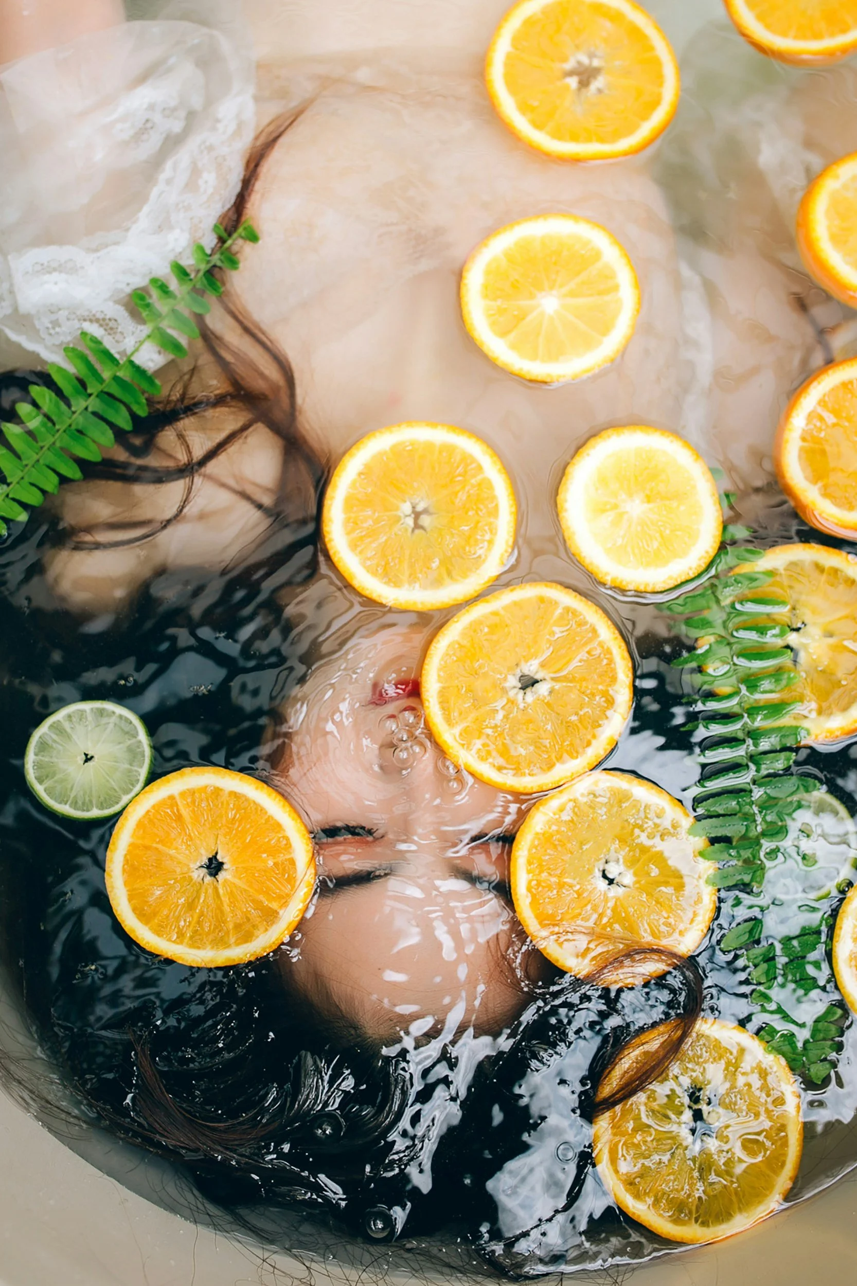 Person submerged in a bath with slices of orange, lemon, and lime, along with green fern leaves and lace fabric.