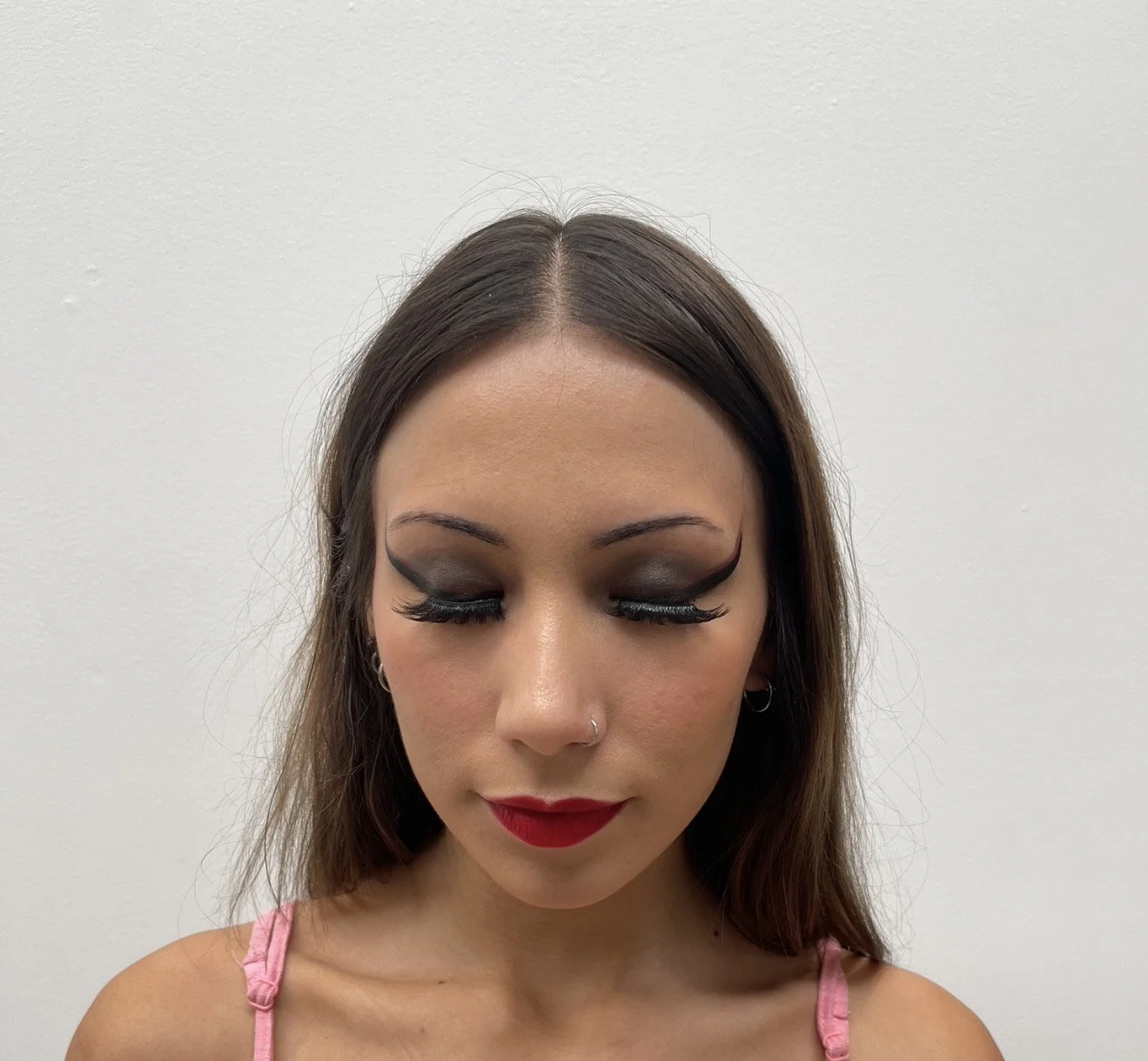 A woman with dark brown hair and a nose ring, wearing pink straps, has her eyes closed showing bold smoky eye makeup and long false eyelashes, with bright red lipstick against a plain white background.