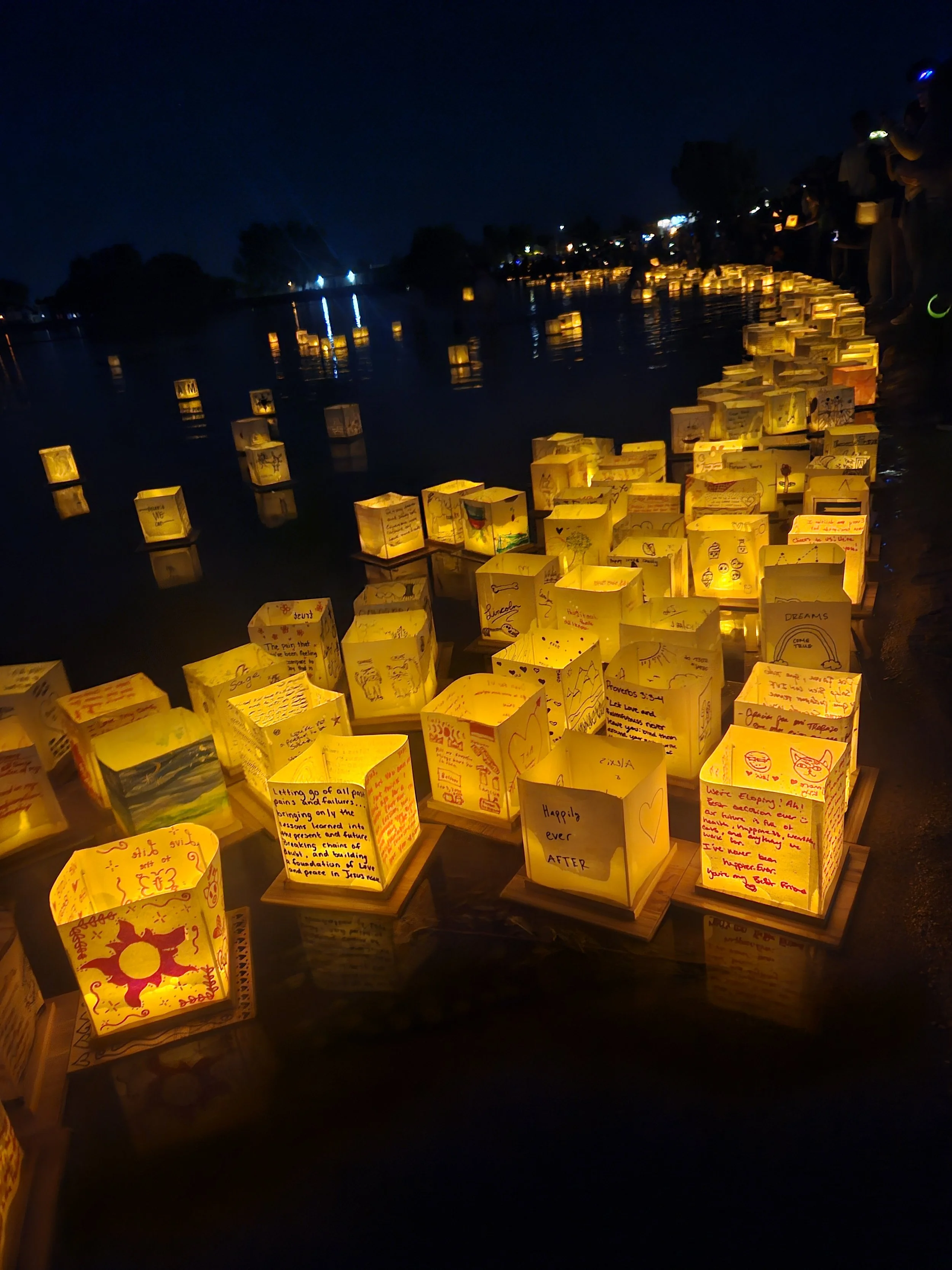 Luminaria paper lanterns lining a waterfront at night, with reflections on the water and people in the background.