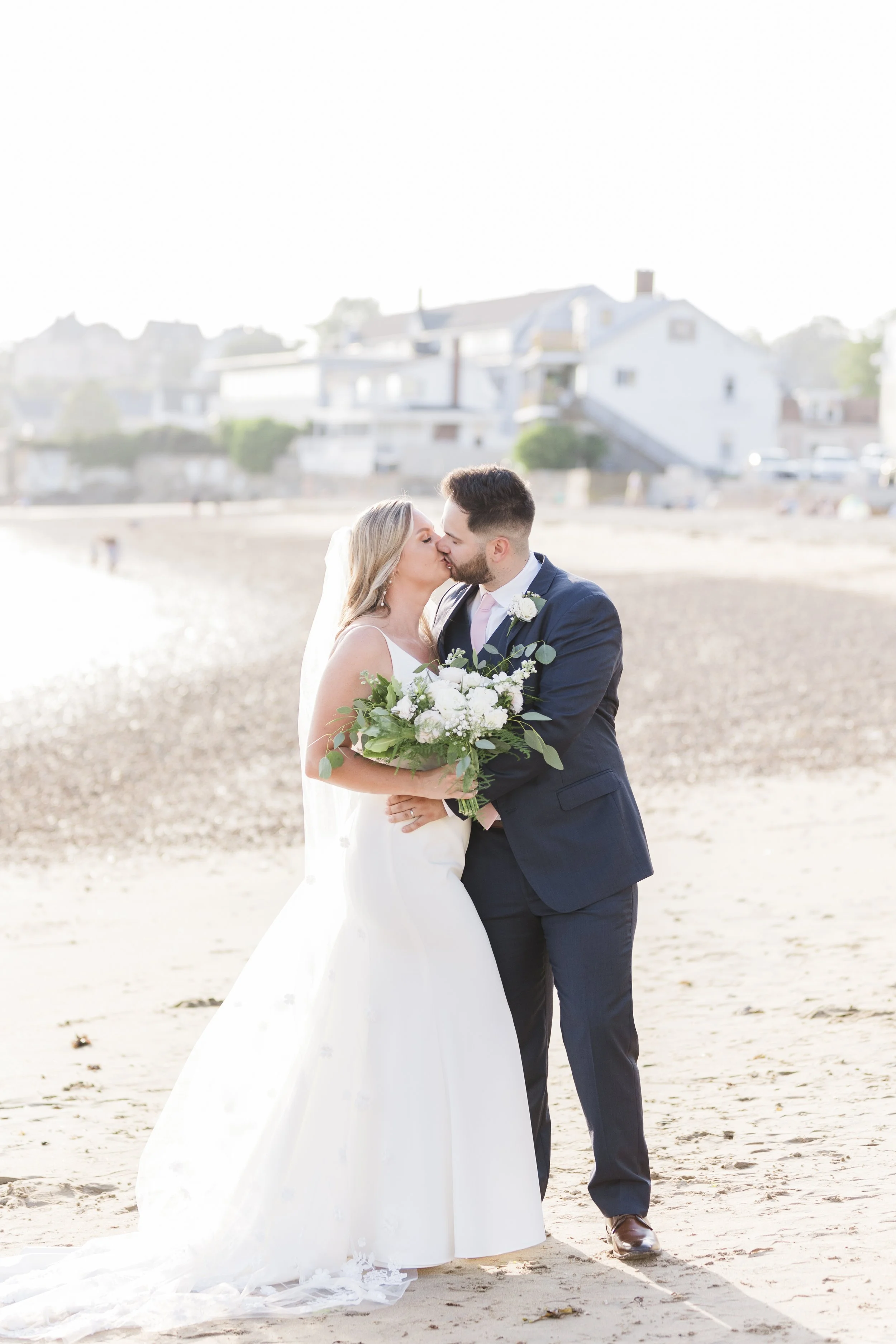A bride and groom share a kiss on a beach during their wedding, with the bride holding a bouquet of white and green flowers and wearing a white dress, while the groom wears a dark suit.