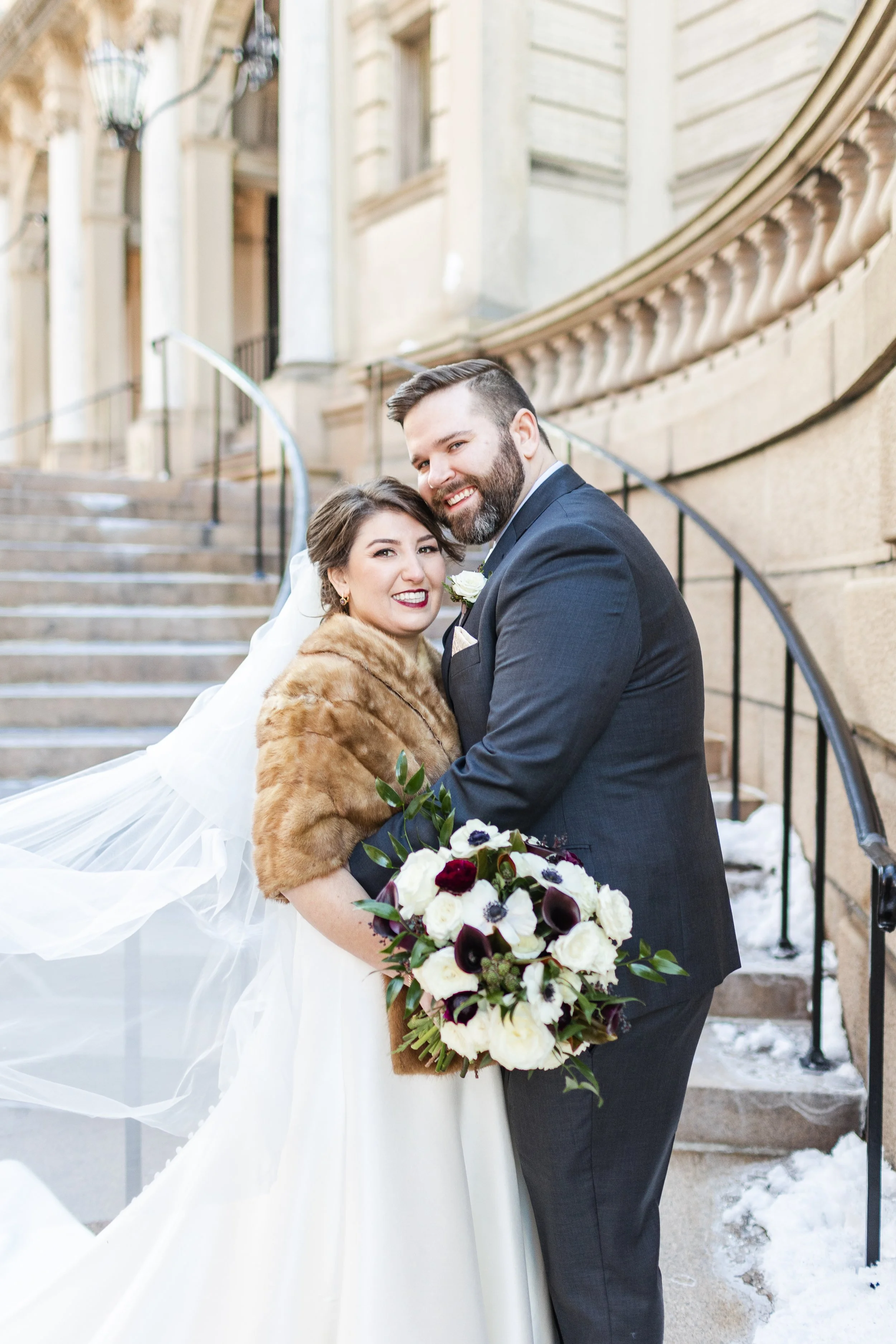 A bride and groom smiling and embracing outdoors on a staircase, bride holding a bouquet of white and dark flowers, winter setting with snow on the ground.