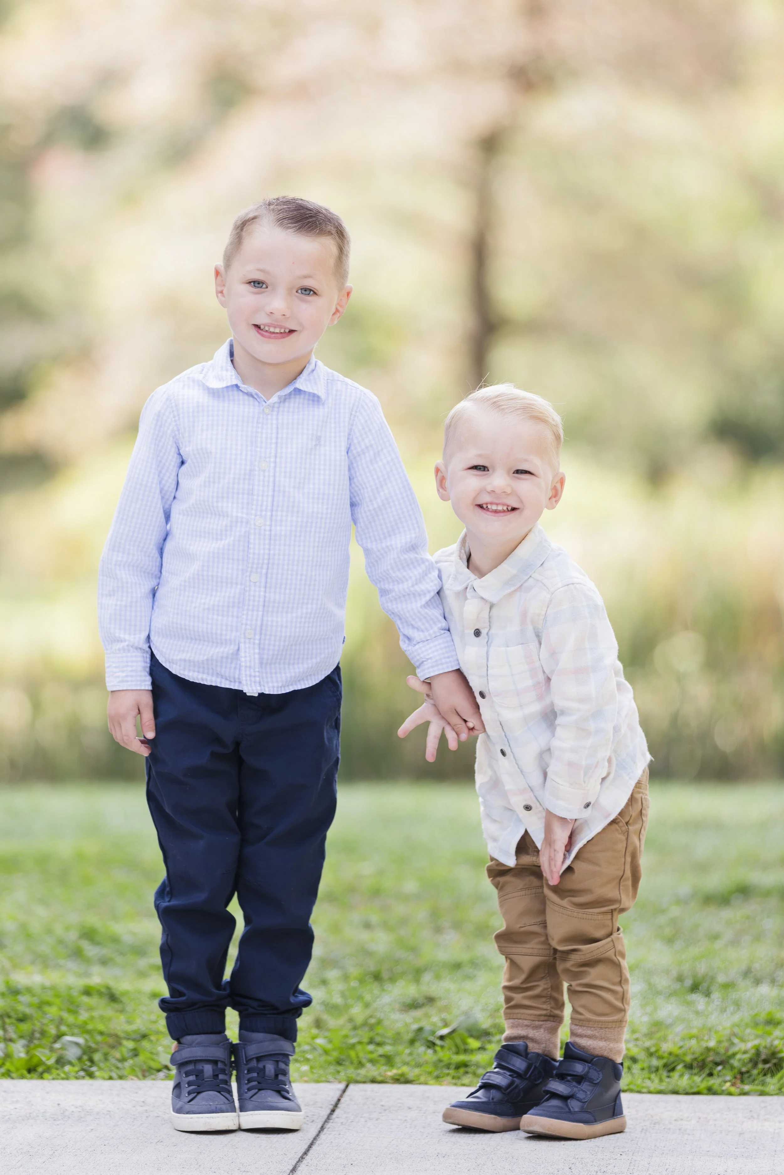 Two young boys smiling and holding hands outdoors on a grassy area with trees in the background, dressed in casual button-up shirts and pants.