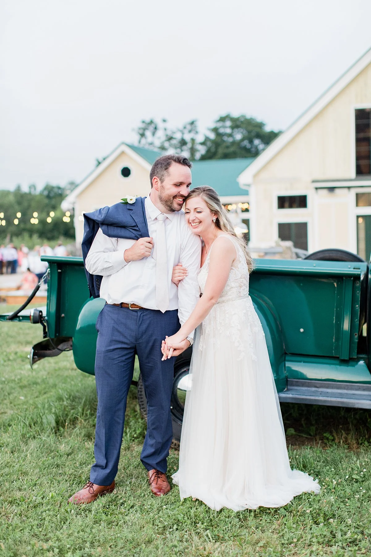 A newlywed couple smiling and laughing outdoors in front of a green vintage truck at their wedding reception. The bride is in a white wedding dress, and the groom is in a white shirt and blue pants, with a jacket draped over his shoulder.