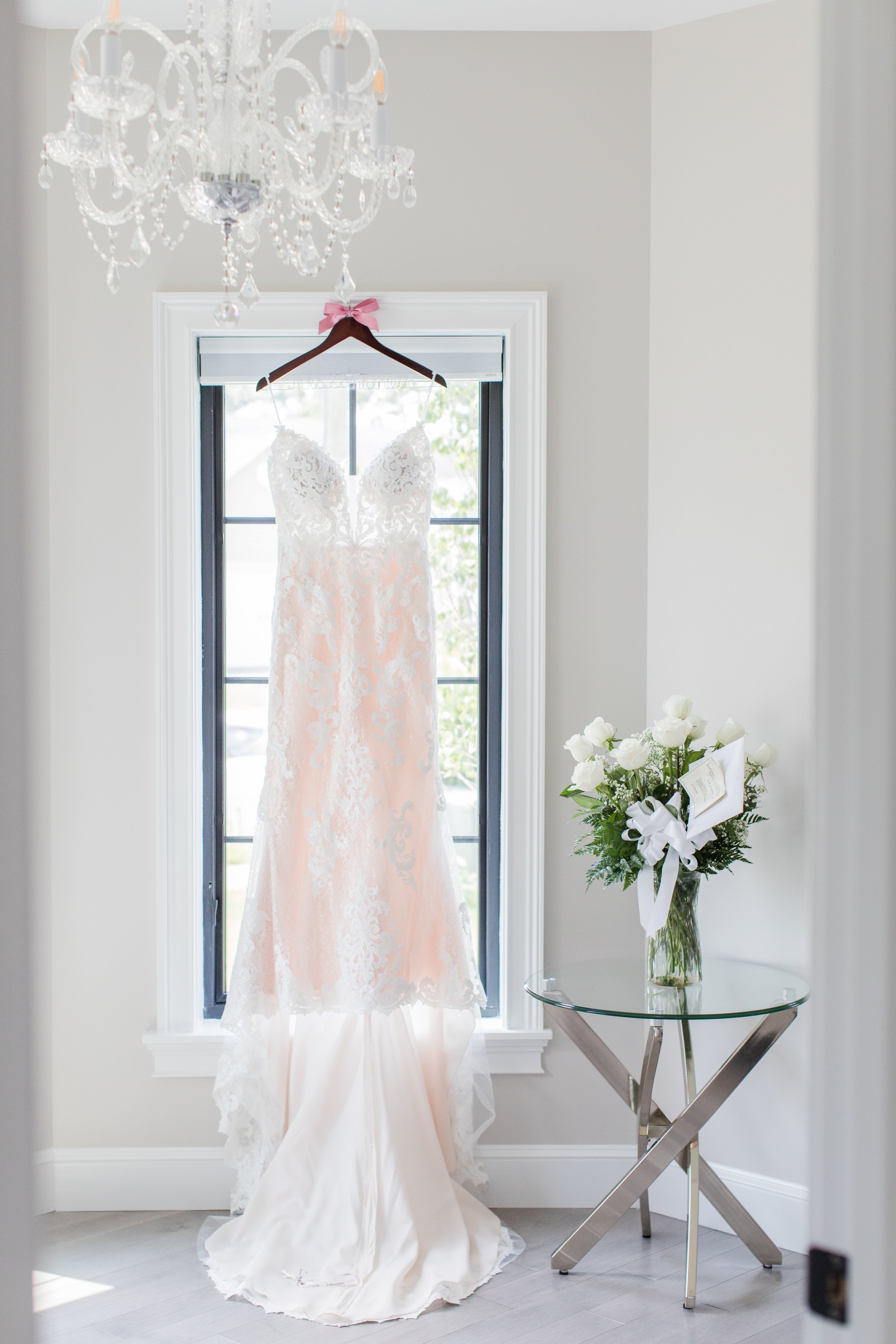 A wedding dress hanging on a hanger with a pink bow, in front of a window in a bright room. A crystal chandelier is above, and a glass side table with a white rose bouquet is to the right.
