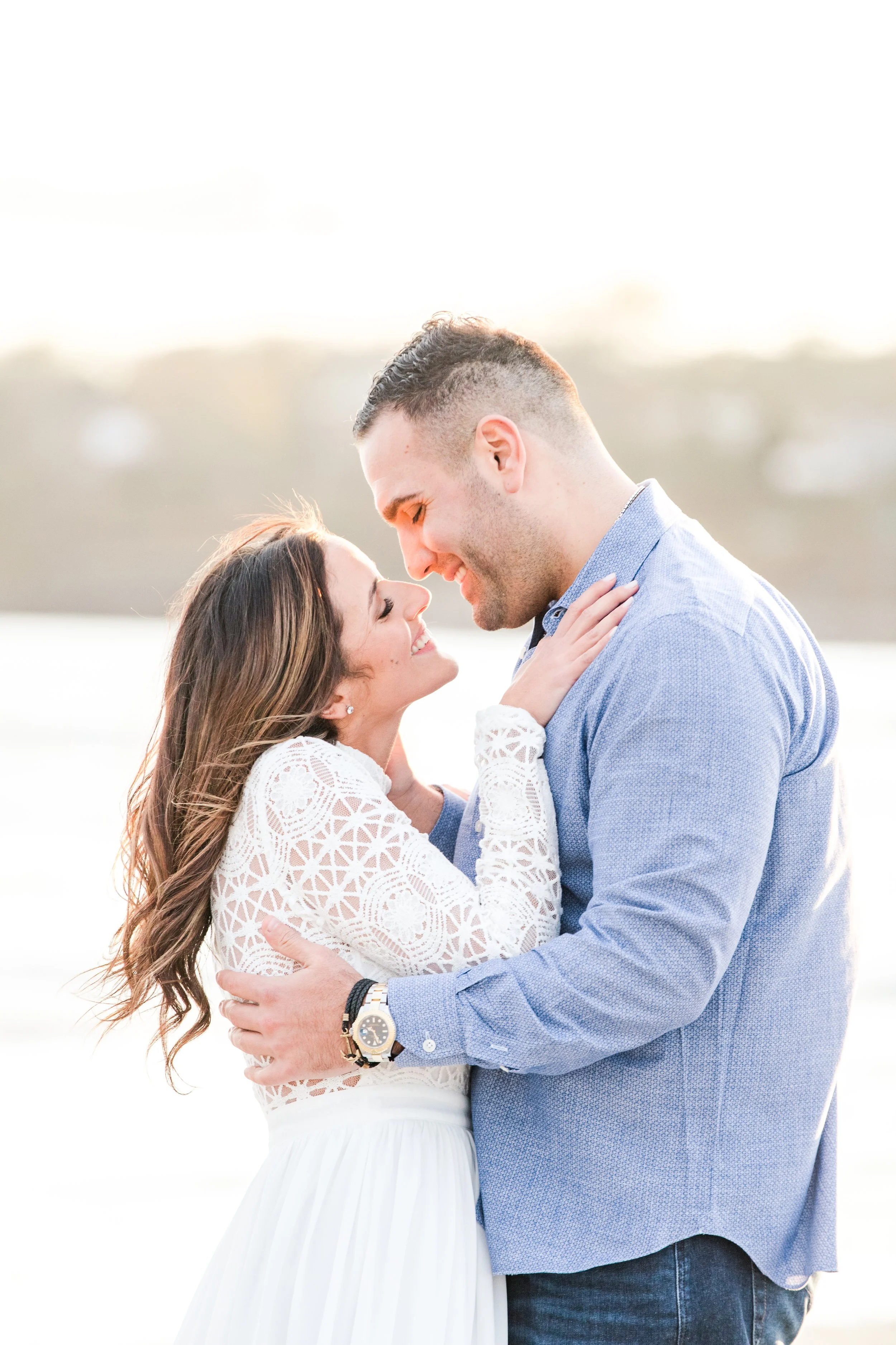 A young couple smiling and embracing outdoors during sunset, with a blurred background of a body of water and distant land.