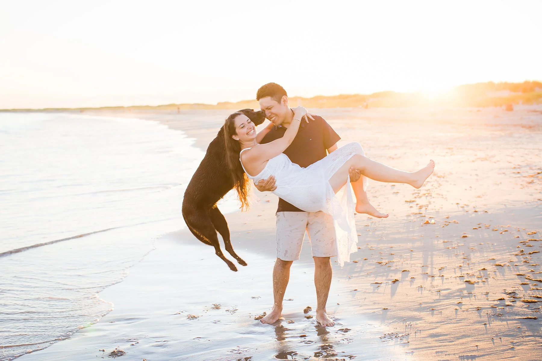 A happy couple on the beach at sunset, with the man holding a woman in a white dress and a black dog jumping up to them.