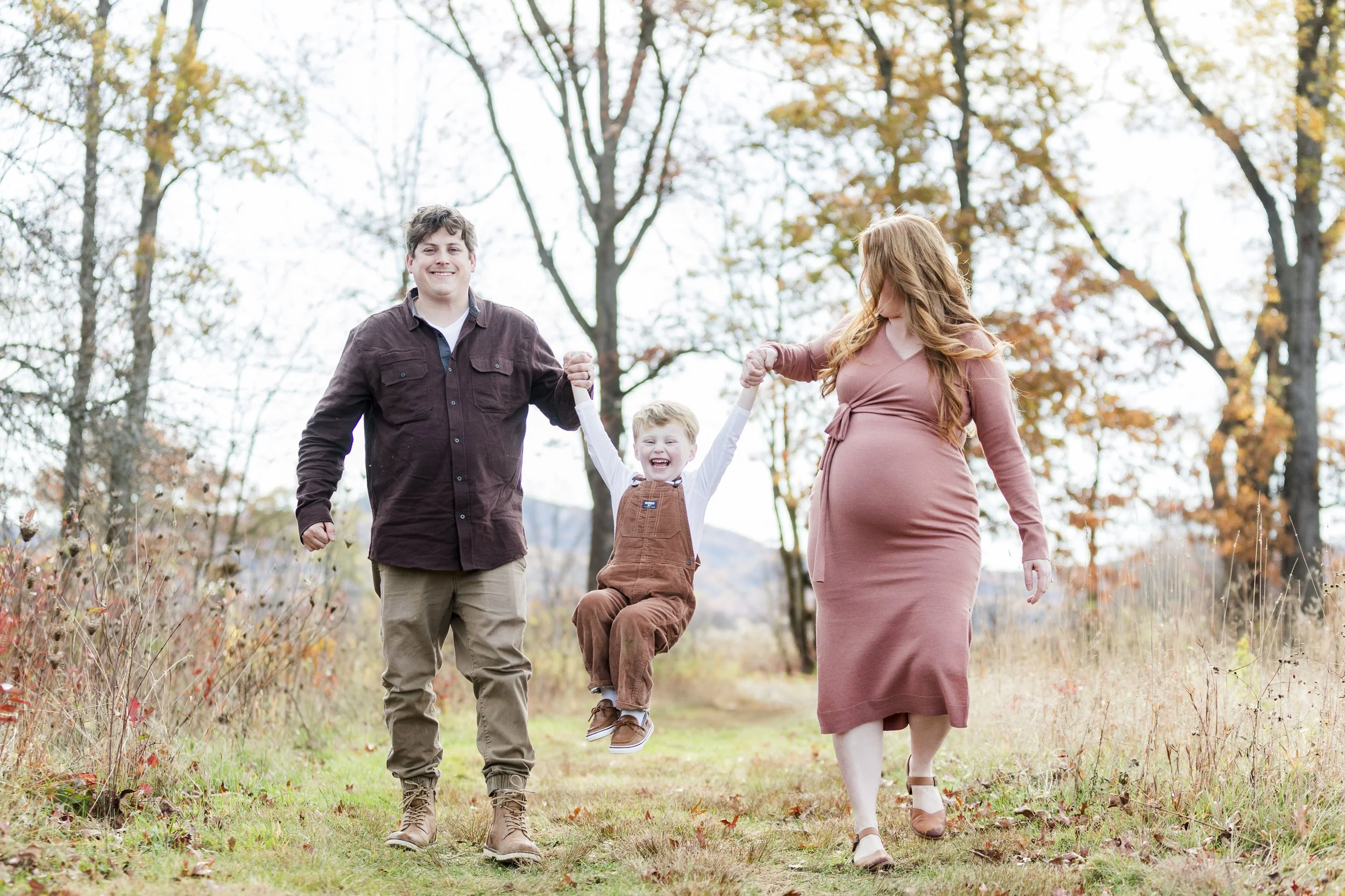 A family of three walking outdoors in an autumn park, with the mother and father holding the child's hands as he jumps smiling.