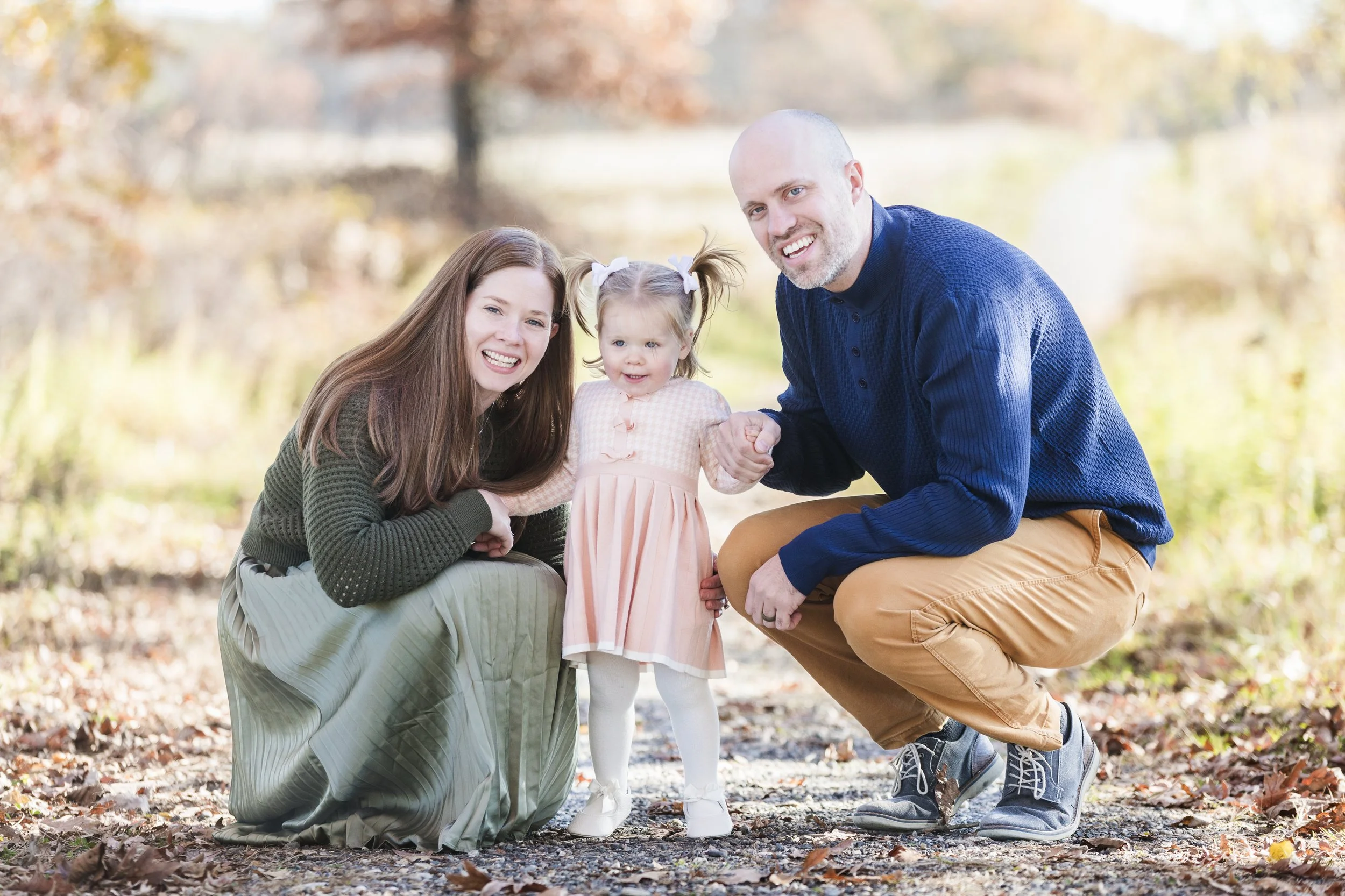 A happy family of three outdoors on a fall day, with autumn leaves on the ground and trees in the background. The mother and father are squatting and holding their young daughter between them, all smiling at the camera.