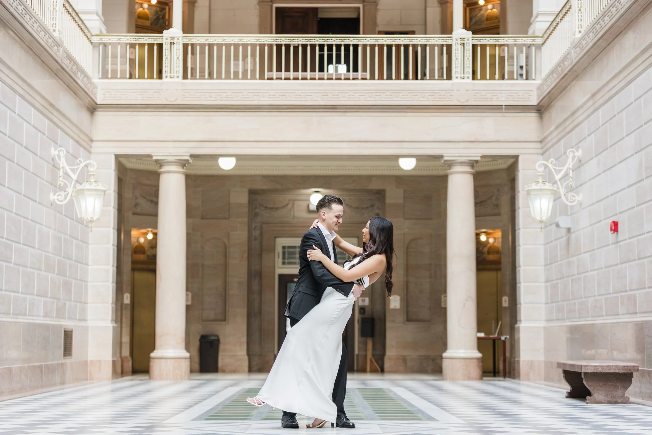 A couple, dressed in wedding attire, dancing and smiling in an elegant, grand indoor setting with high ceilings, columns, and decorative wall-mounted lamps.