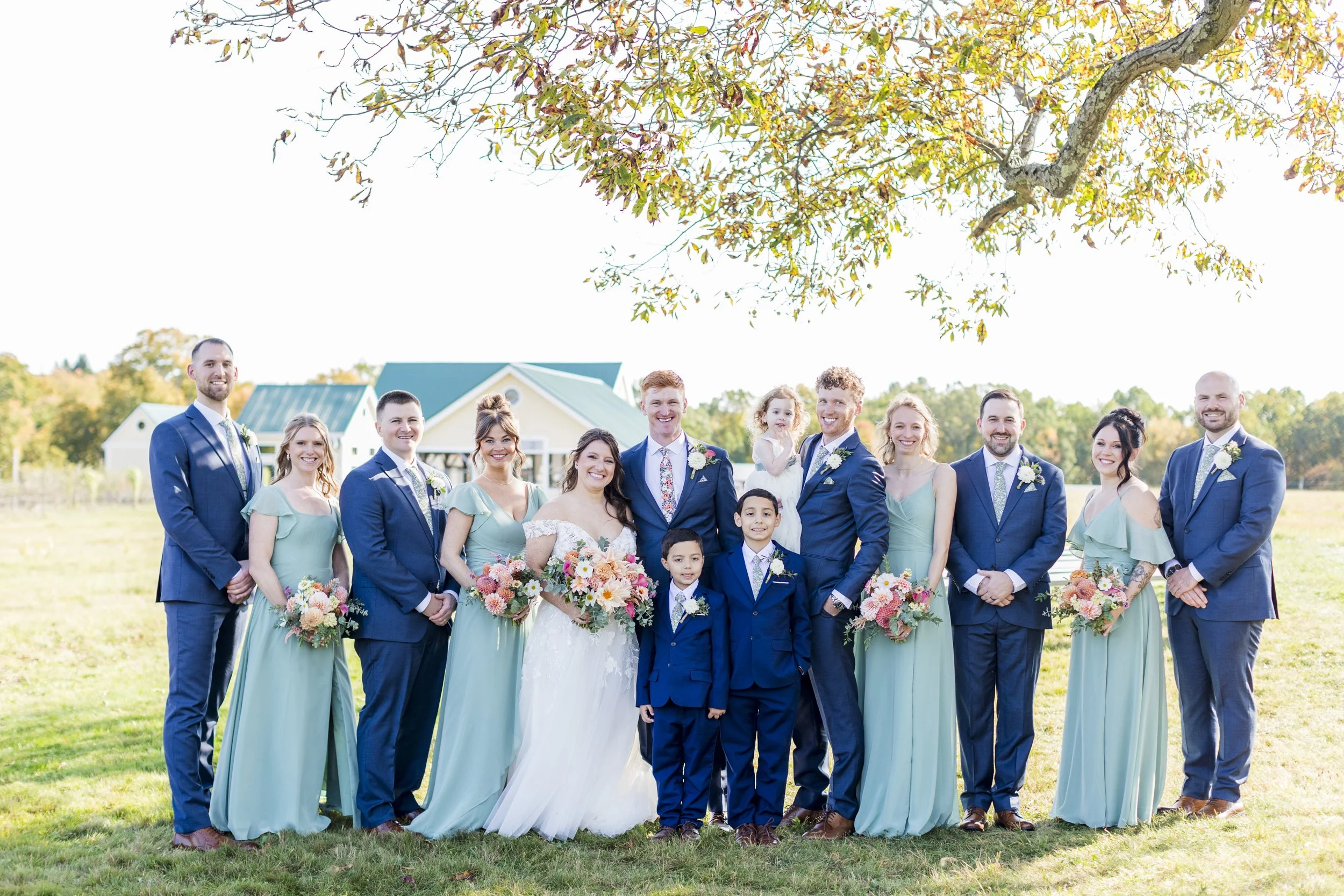 A wedding party of fifteen people, including a bride, groom, bridesmaids, groomsmen, and children, standing outdoors on grass with trees and a small building in the background, during daytime with clear weather.