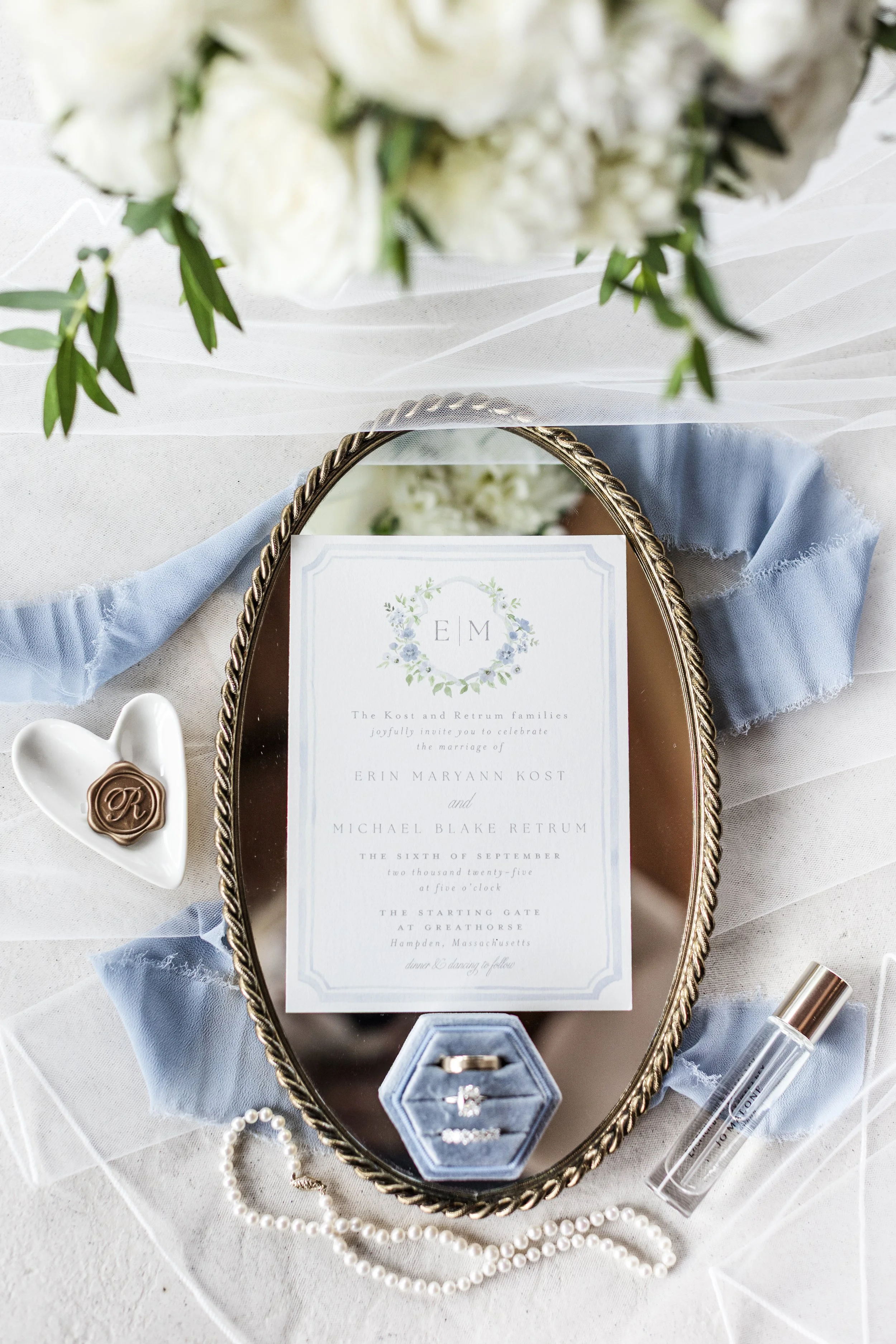 A wedding invitation display with a mirror, ring box, pearl necklace, small heart dish, and wedding bouquet above