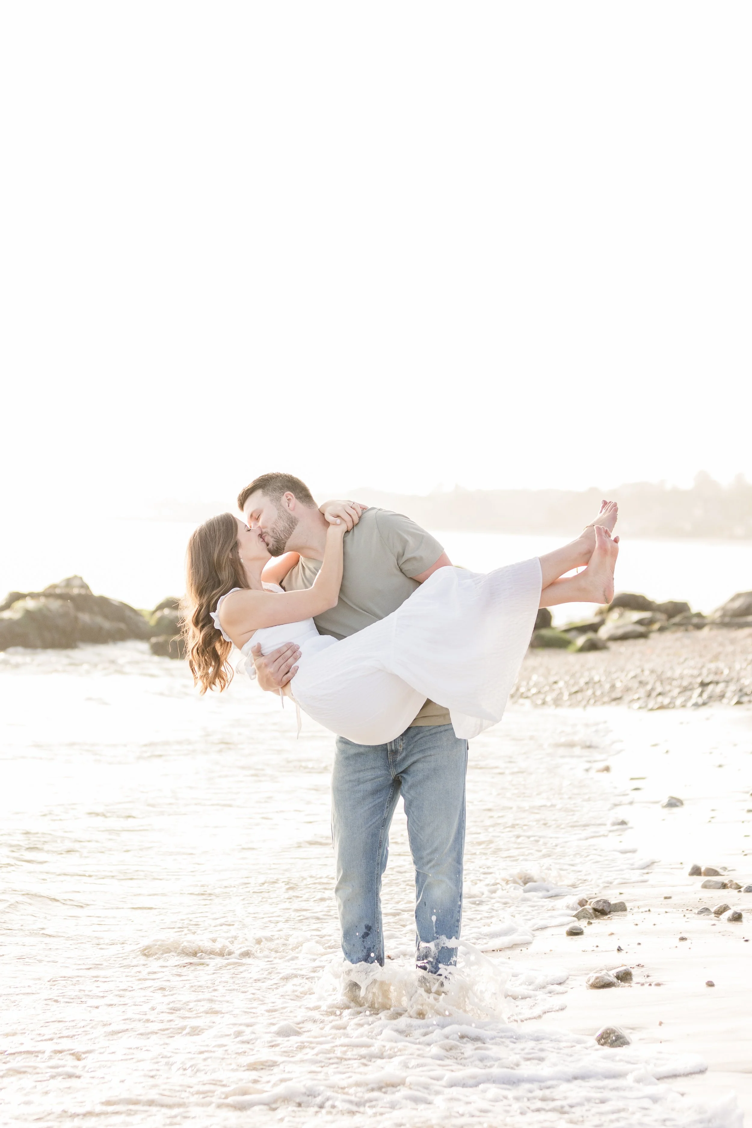 A man is holding a woman in his arms, and they are kissing on a beach at sunset. The woman has long brown hair and is wearing a white dress, while the man has short hair and a beard, wearing a gray t-shirt and jeans. Waves are splashing around their 