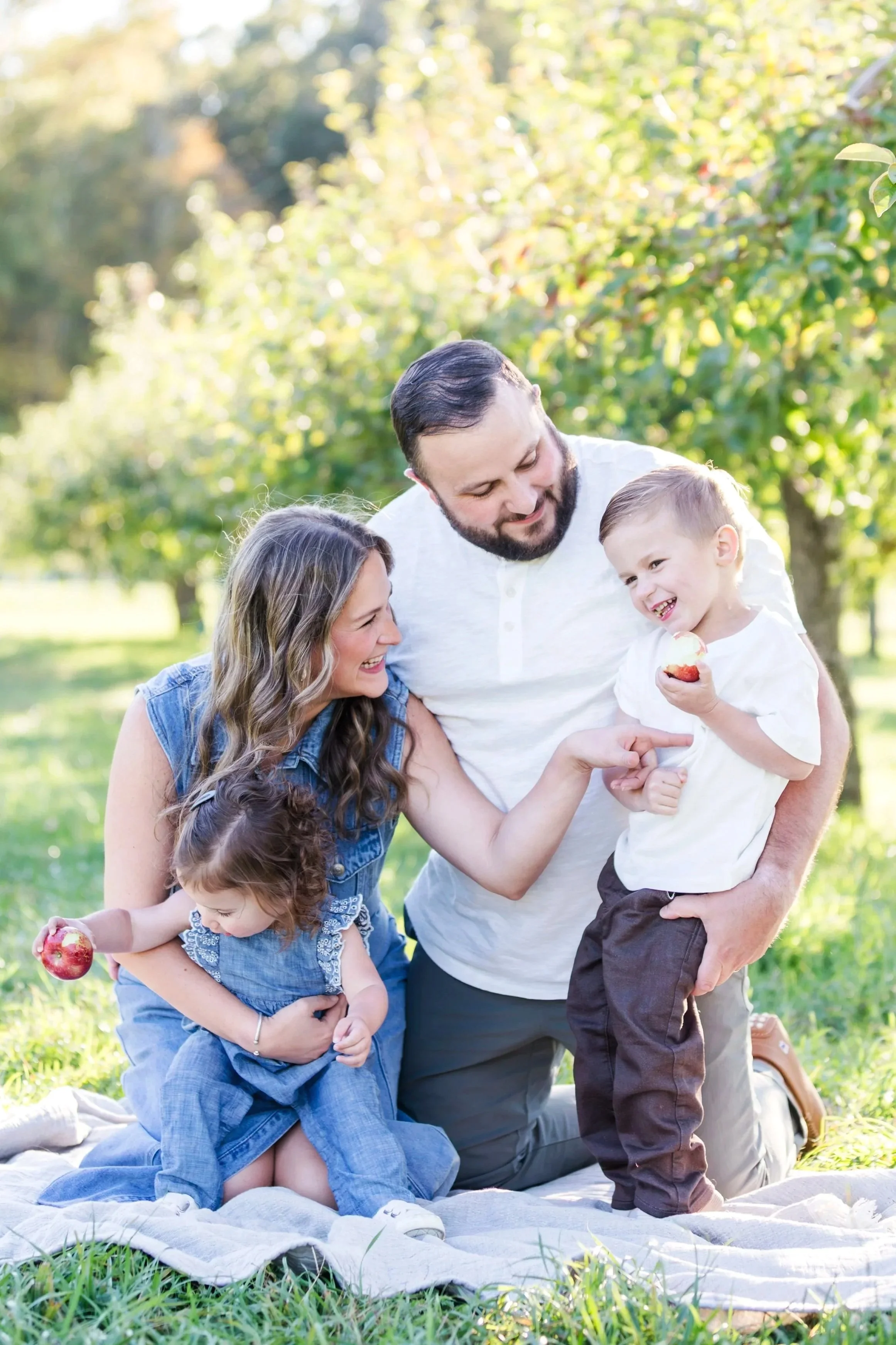 Family enjoying a picnic outdoors, sitting on a blanket on the grass, with trees and sunlight in the background. The mother, father, and two children are smiling and sharing snacks.