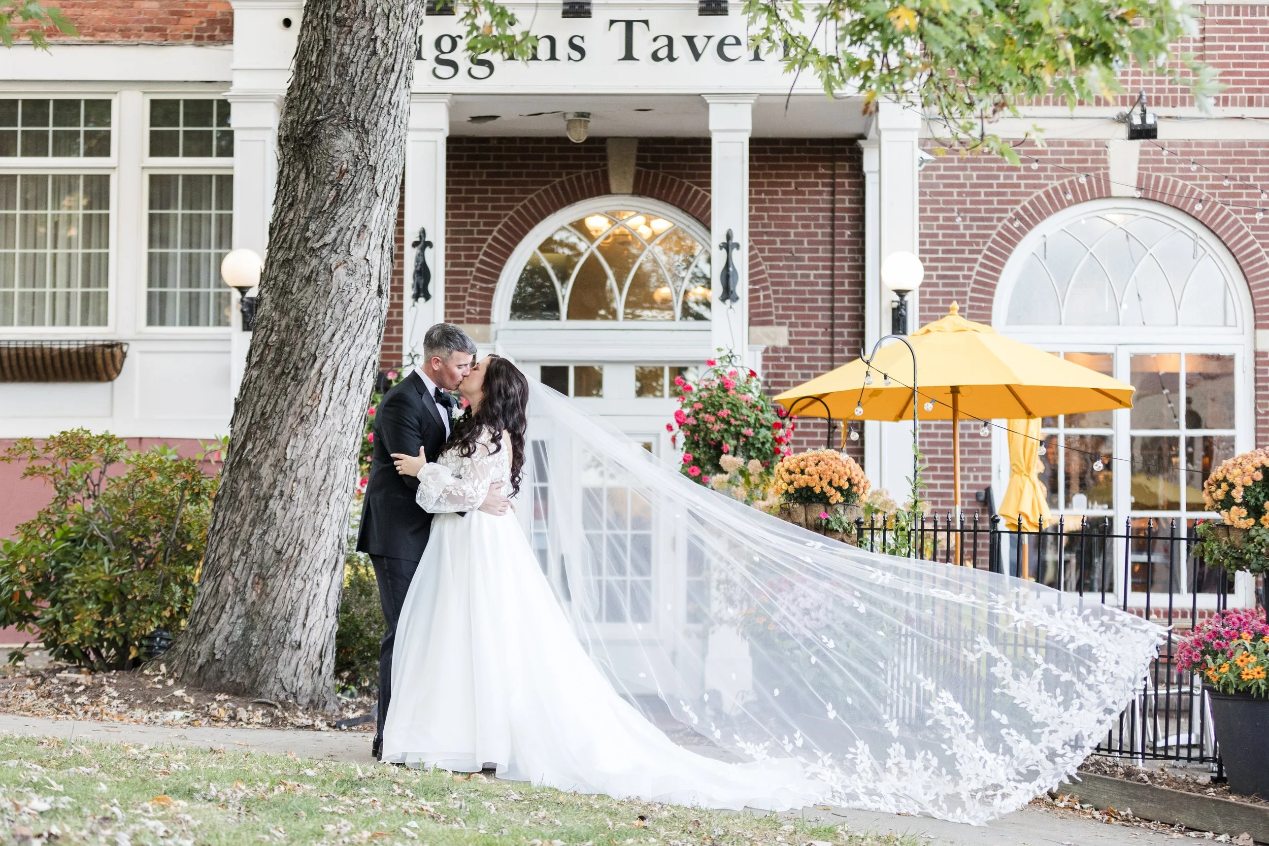 A bride and groom share a kiss outside a brick building with a sign that says 'Stiggins Tavern.' The bride wears a white wedding gown with a long train and a veil, while the groom is dressed in a black tuxedo. The scene includes colorful flower pots,